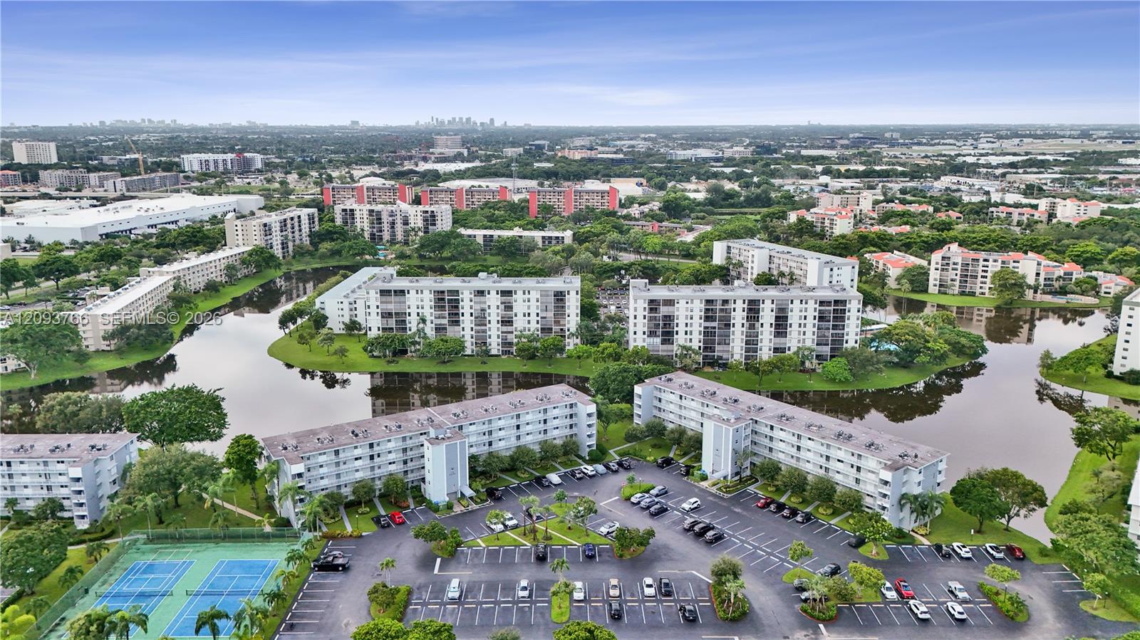 Aerial view of white condominium buildings around a lake, with tennis courts and parking below.