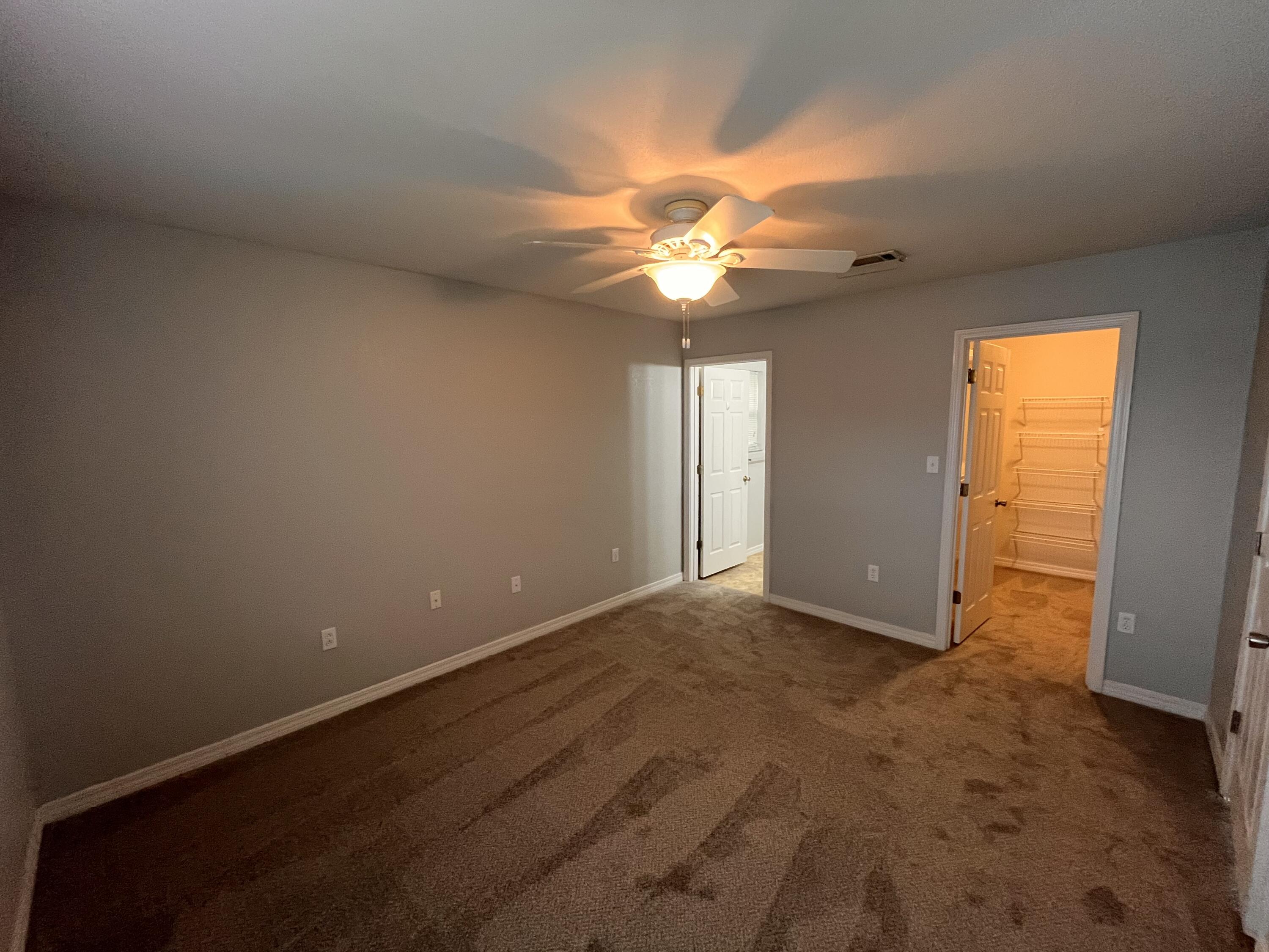 Empty carpeted bedroom with a lit ceiling fan and an open closet with wire shelving.