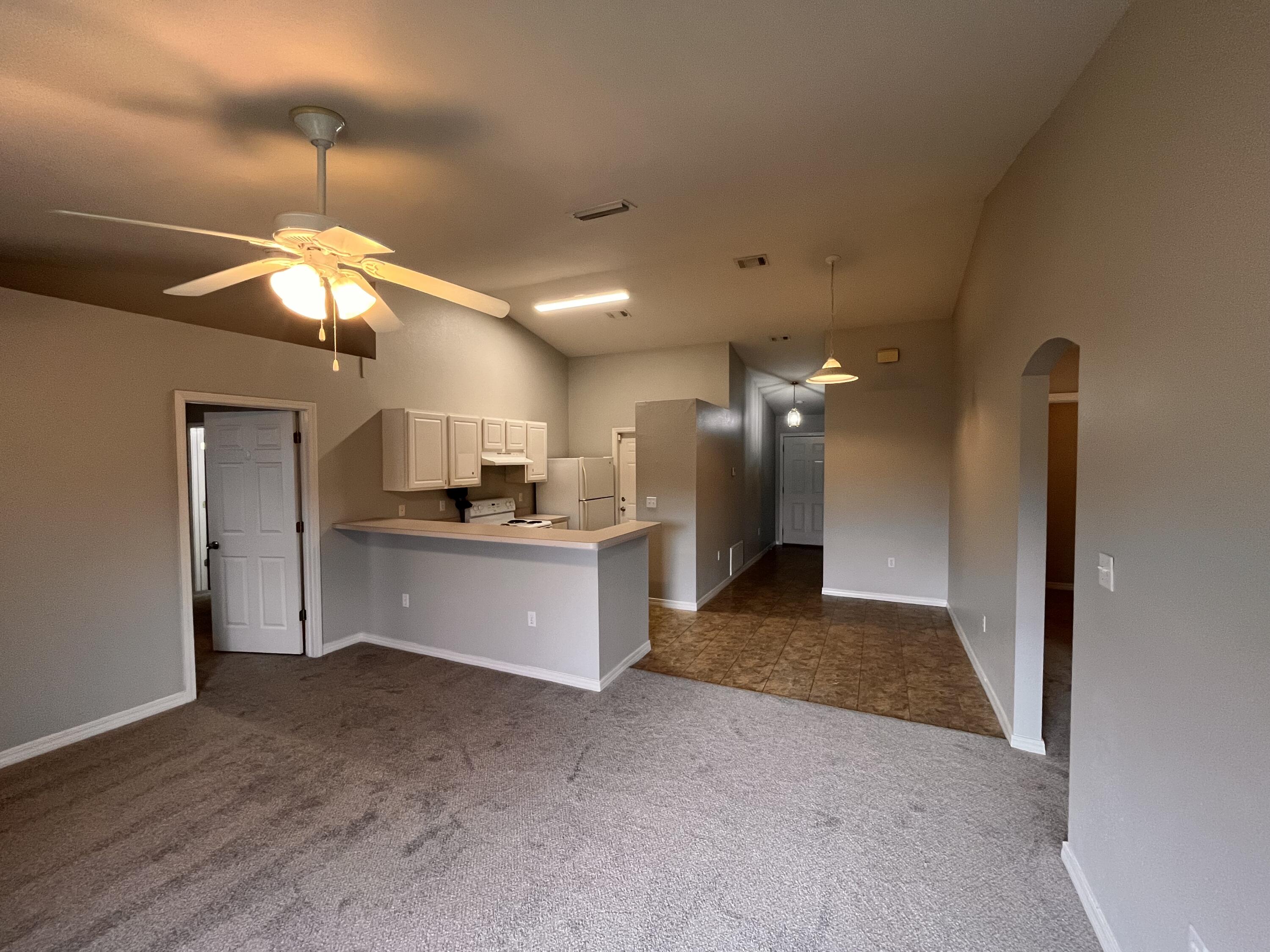Vacant living room with gray carpet, ceiling fan, and open kitchen with breakfast bar.