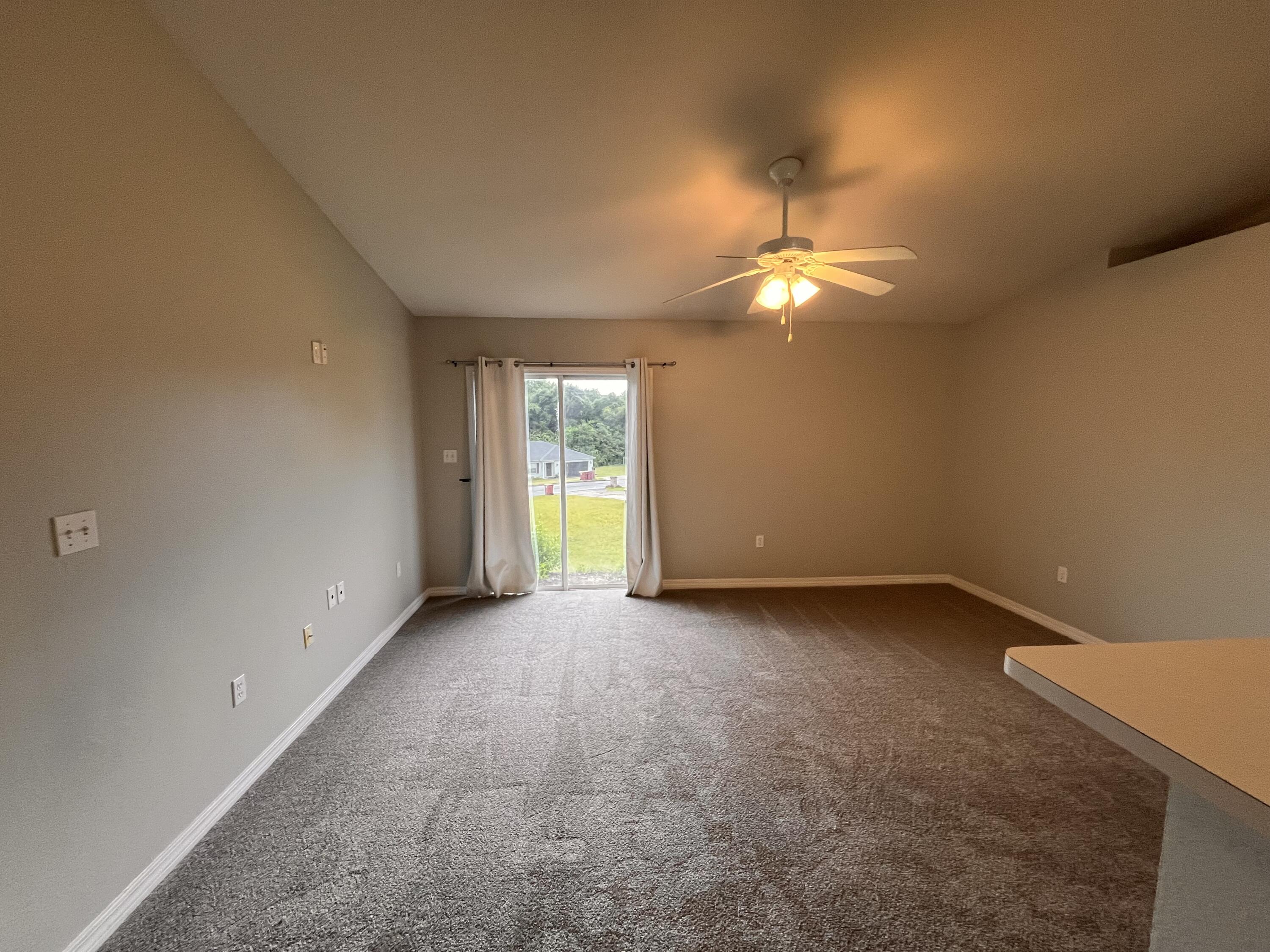 Empty carpeted bedroom with a ceiling fan and curtained sliding glass door overlooking a lawn.