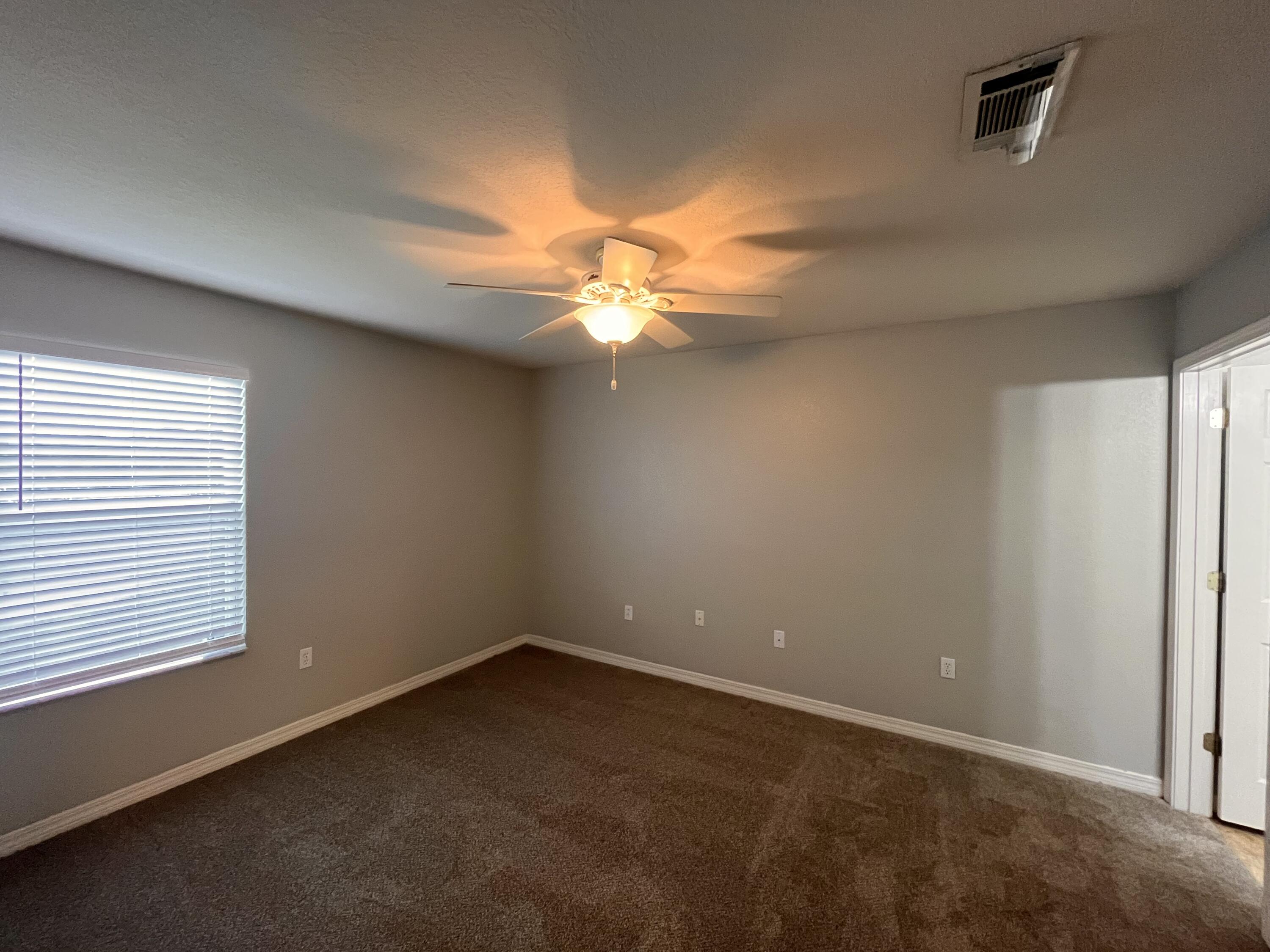 Empty carpeted room with gray walls, window blinds, and a ceiling fan with its light on.