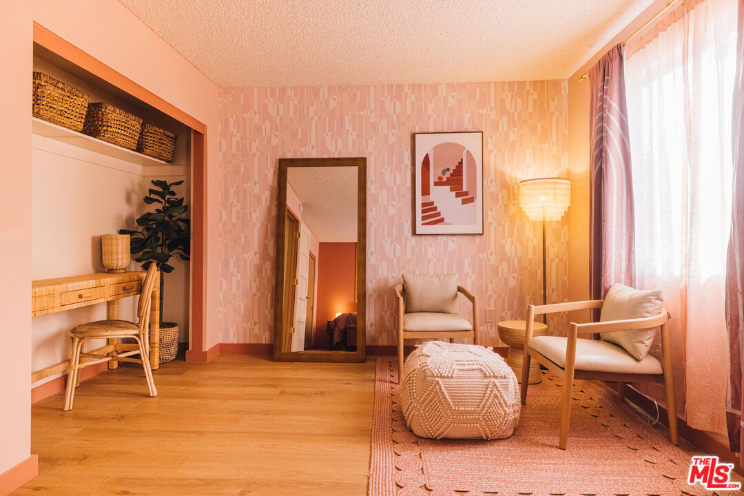Pink living room with patterned wallpaper, two chairs, a knitted pouf, and a tall mirror.