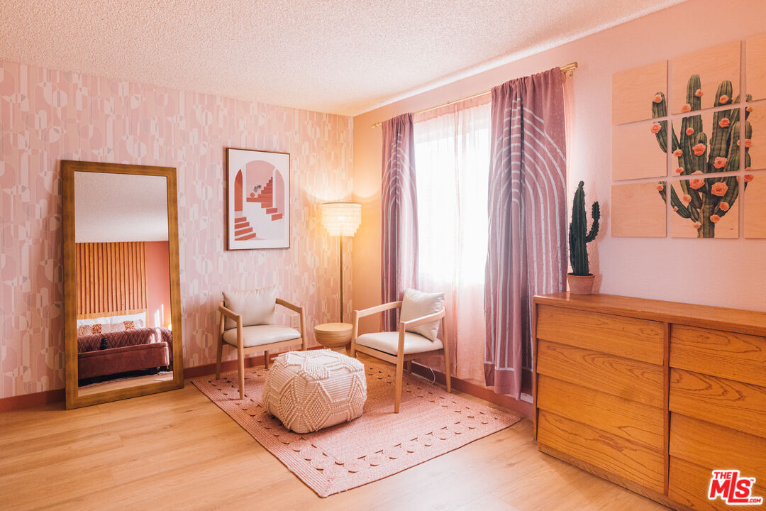 Furnished bedroom sitting area with blush wallpaper, a pink rug, wooden dresser, and cactus artwork.
