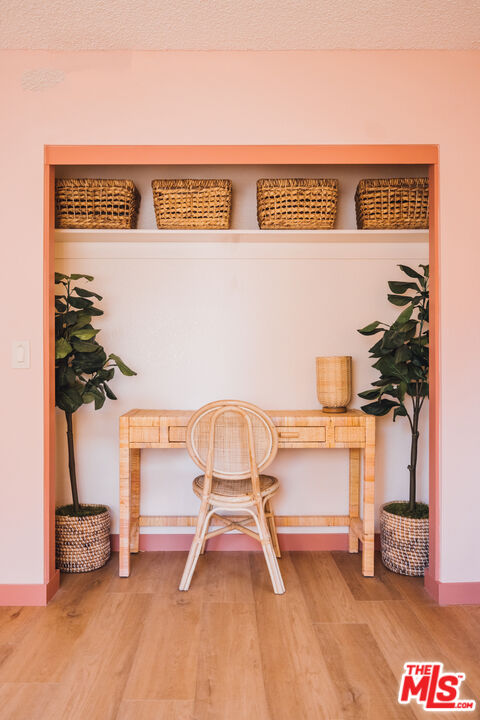 Rattan desk nook framed by coral-pink walls, with a woven chair, shelf baskets, and potted plants.