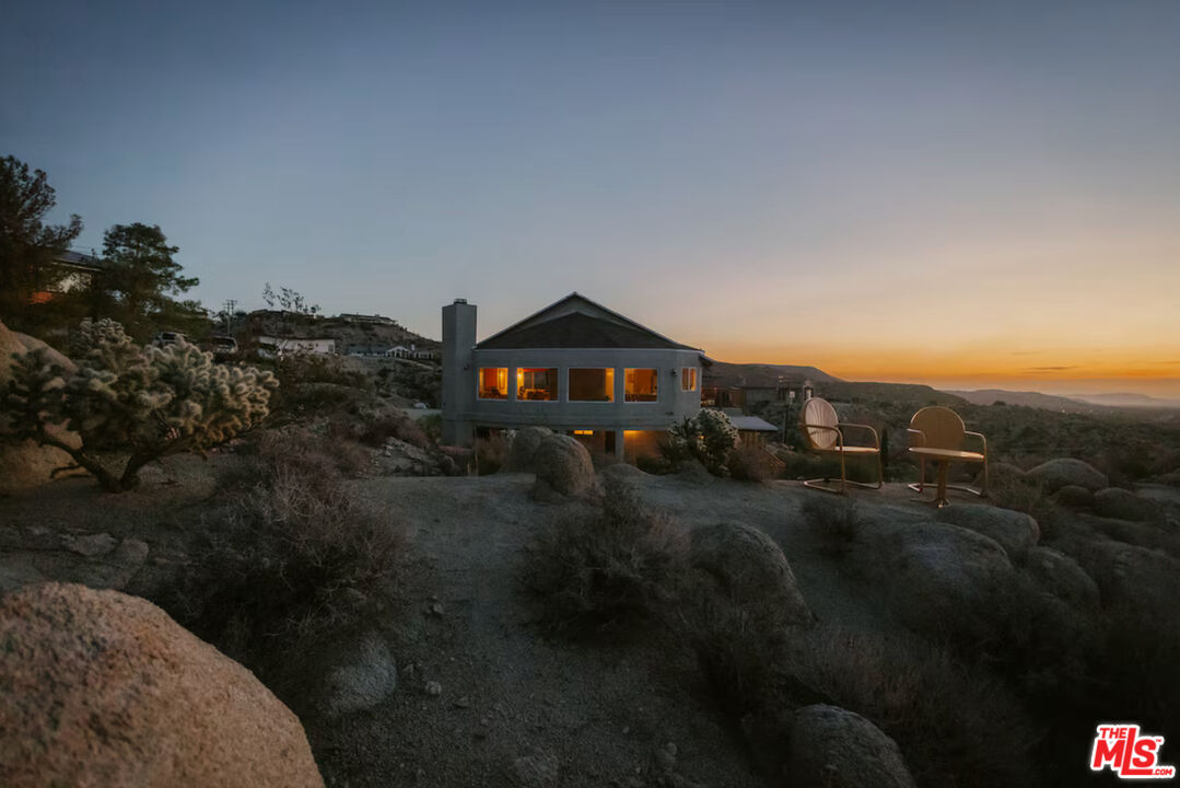Modern gray home among boulders, with glowing windows and two chairs overlooking hills at sunset.
