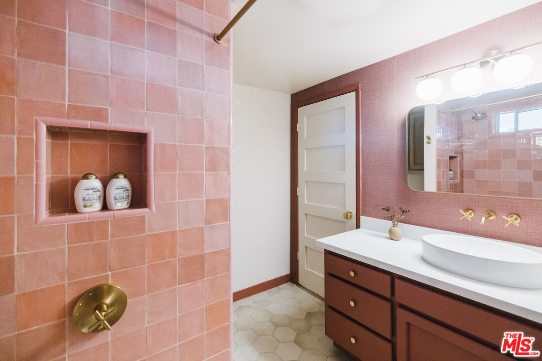 Bathroom with pink tiled shower, brass fixtures, and a white vessel sink on a reddish-brown vanity.