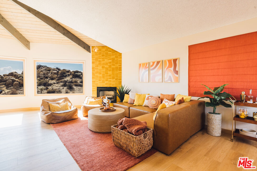 Living room with vaulted wood-beam ceiling, yellow tile fireplace, and windows overlooking rocky desert terrain.