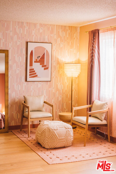 Sitting area with two cream chairs, a textured ottoman, pink patterned wallpaper, and a sunlit window.