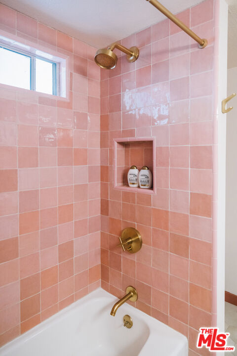 Pink-tiled bathtub and shower with brass fixtures, a recessed shelf, and a small window.