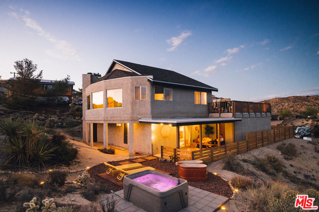 Two-story desert home at dusk with a lit patio, deck, and hot tub.