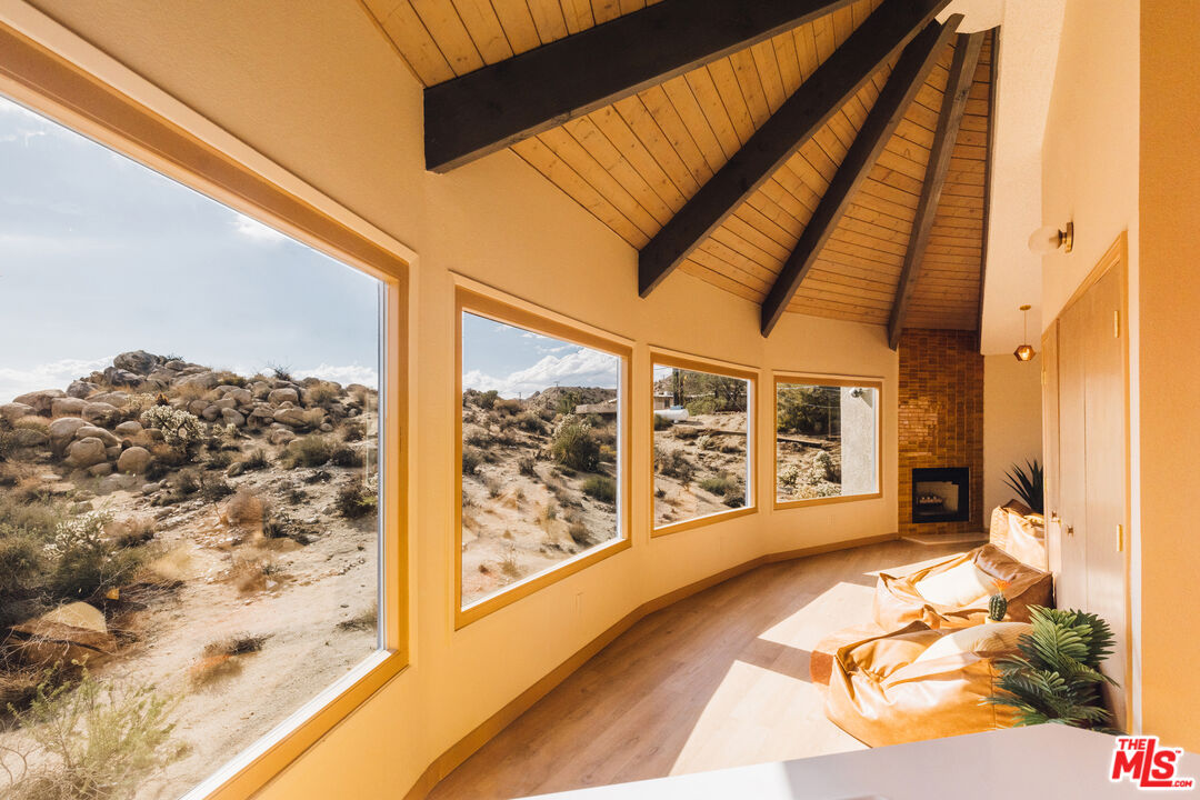 Living room with vaulted wood ceiling, brick fireplace, and large windows overlooking rocky desert hills.