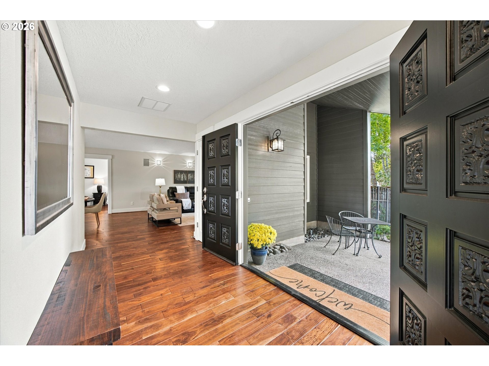 Open black front doors reveal a hardwood entryway and staged living room beside a covered patio.