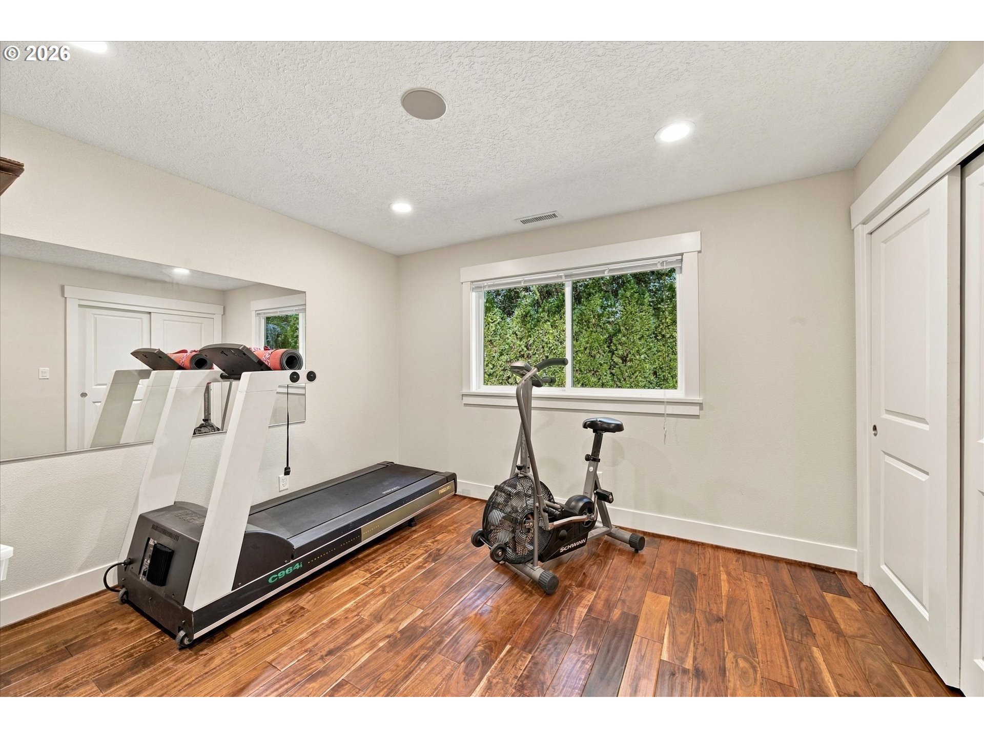 Home gym with a treadmill, air bike, mirrored wall, and window overlooking greenery.