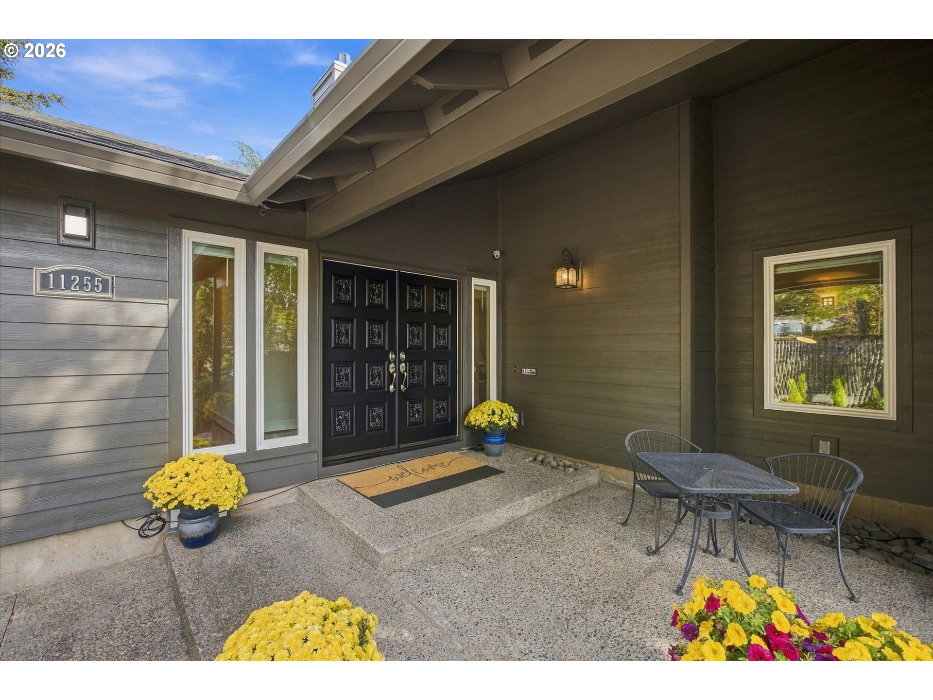 Covered front entry with black double doors, gray siding, patio seating, and yellow flowers.