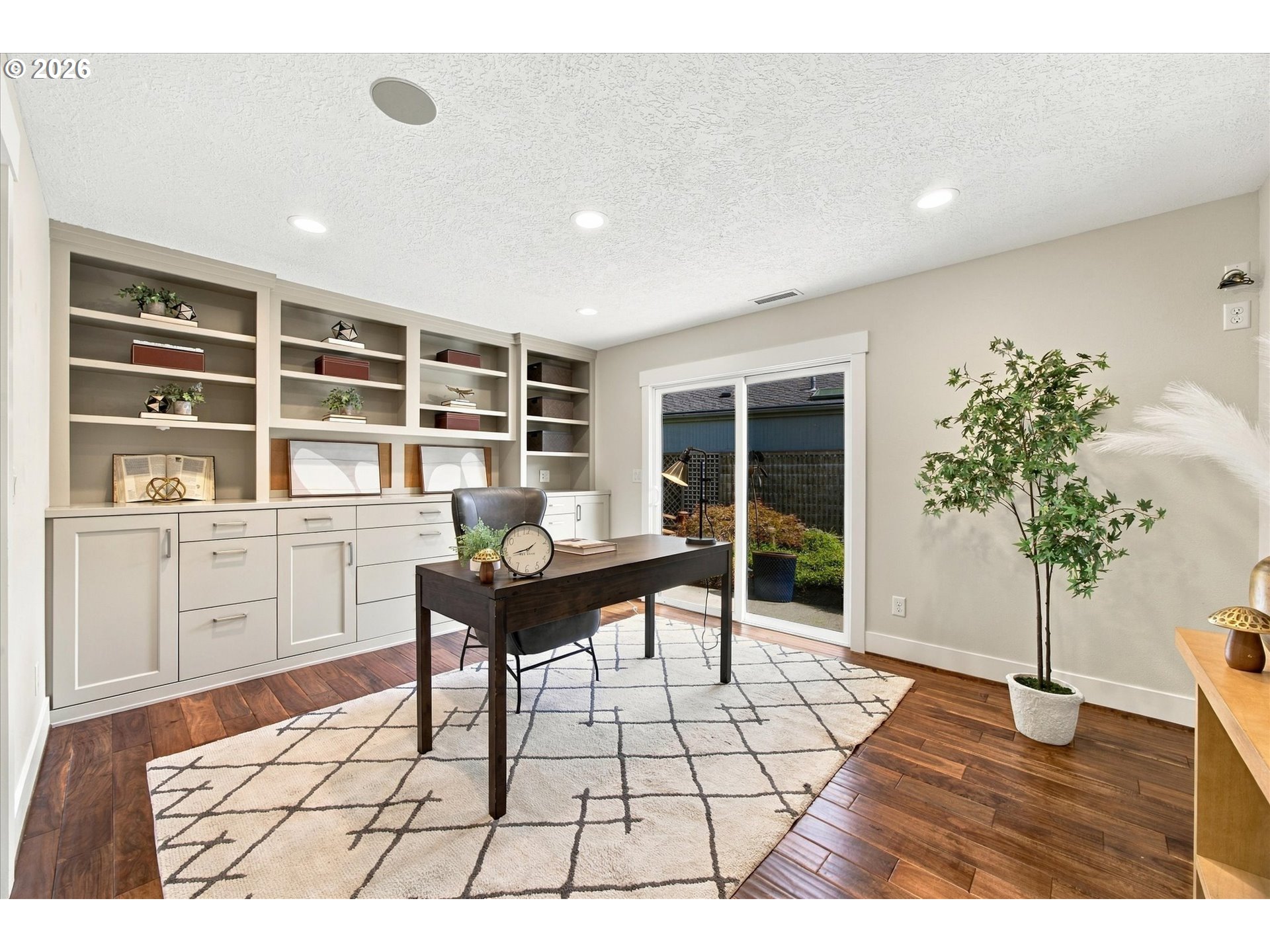 Staged home office with built-in shelves, a dark desk, patterned rug, and garden-facing sliding glass door.