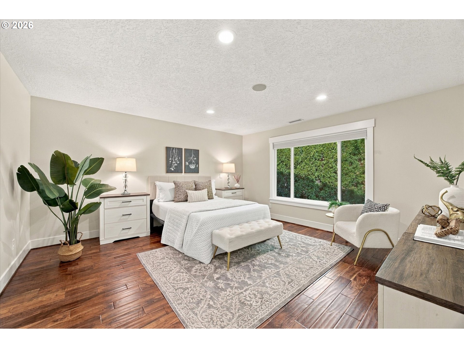 Staged bedroom with a white bed, dark wood floors, and a large window overlooking greenery.