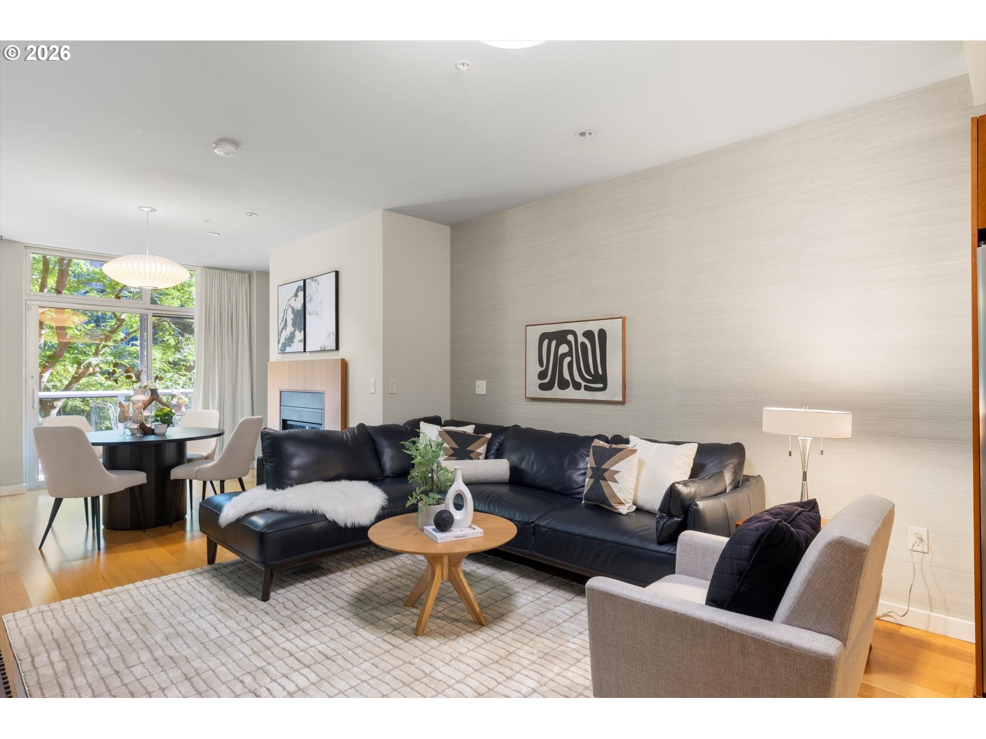 Staged living room with a black leather sectional, round coffee table, and dining area beside large windows.