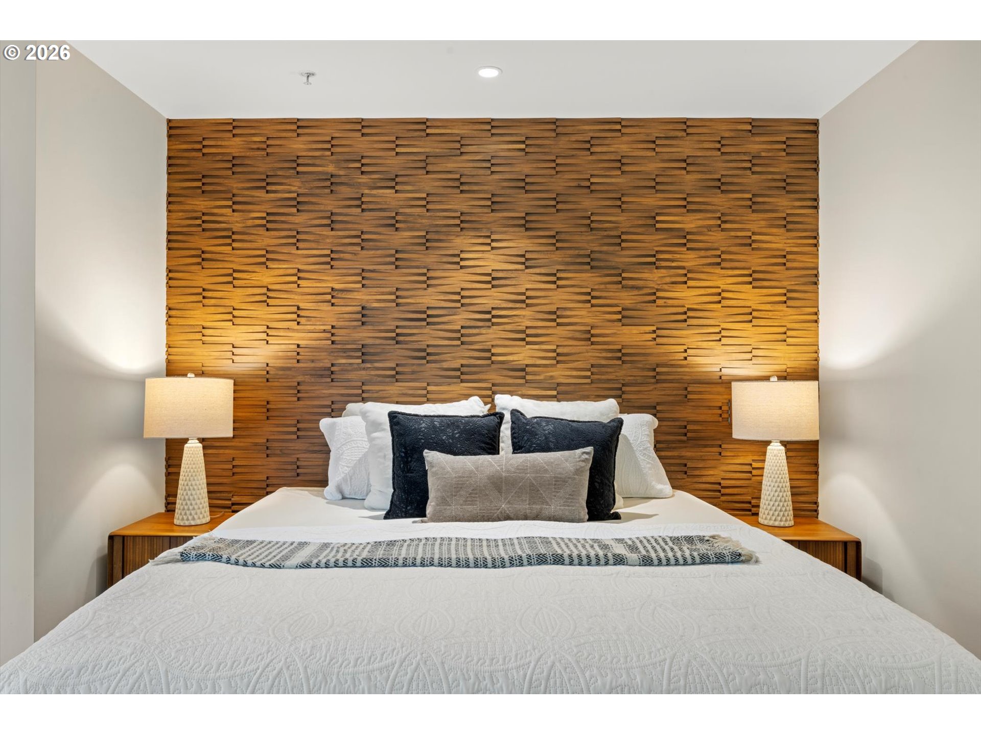 Staged bedroom with a white bed, sculptural wood accent wall, and matching bedside lamps.