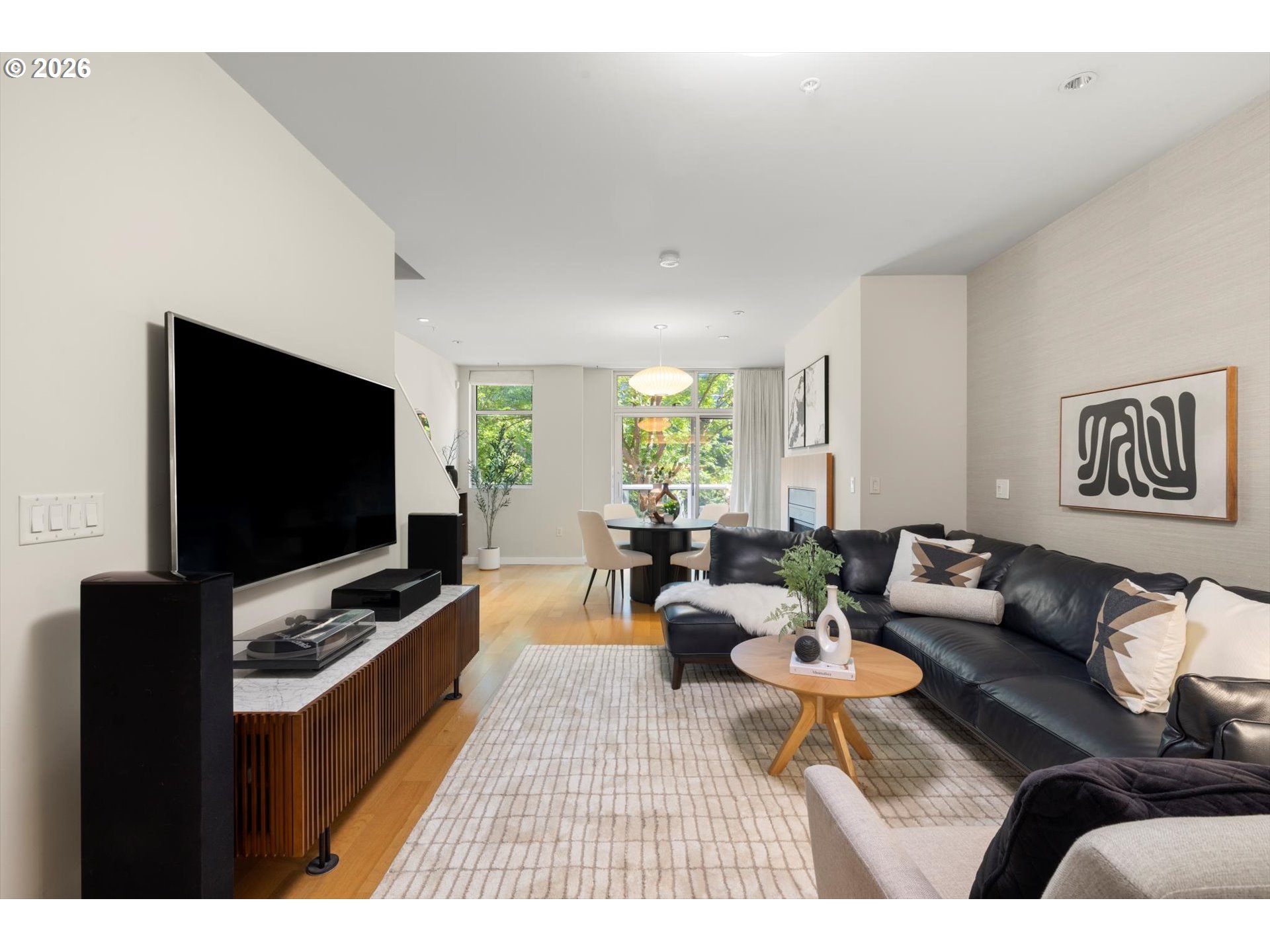 Living room with a black leather sectional, wall-mounted television, and dining area by leafy windows.