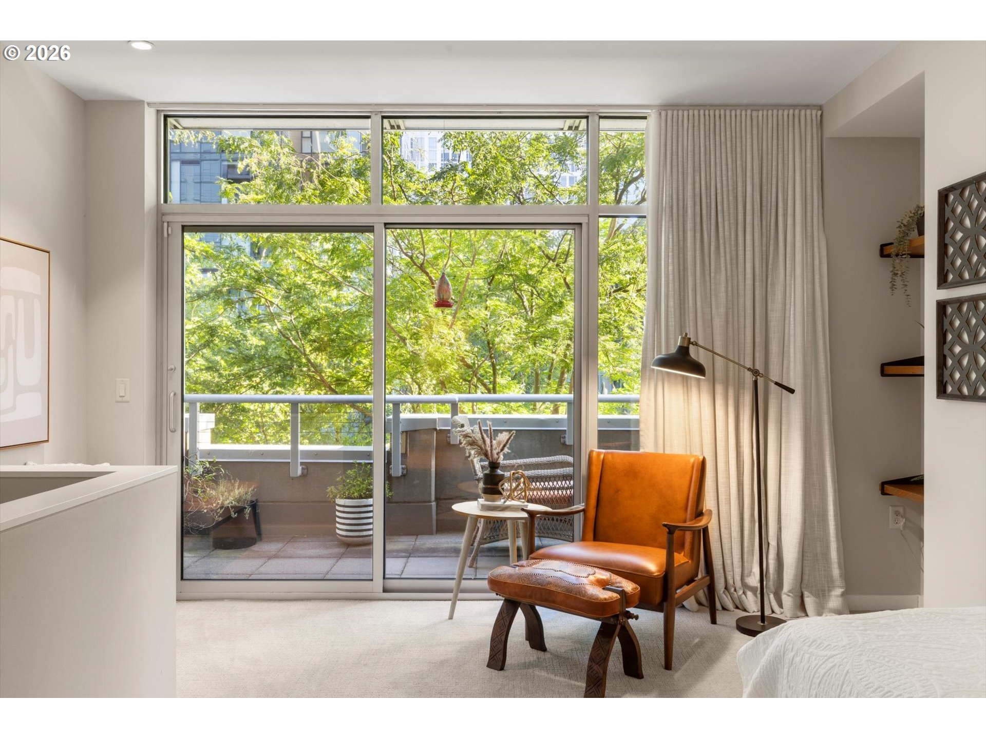 Furnished bedroom with a leather armchair beside tall glass doors opening onto a leafy balcony.