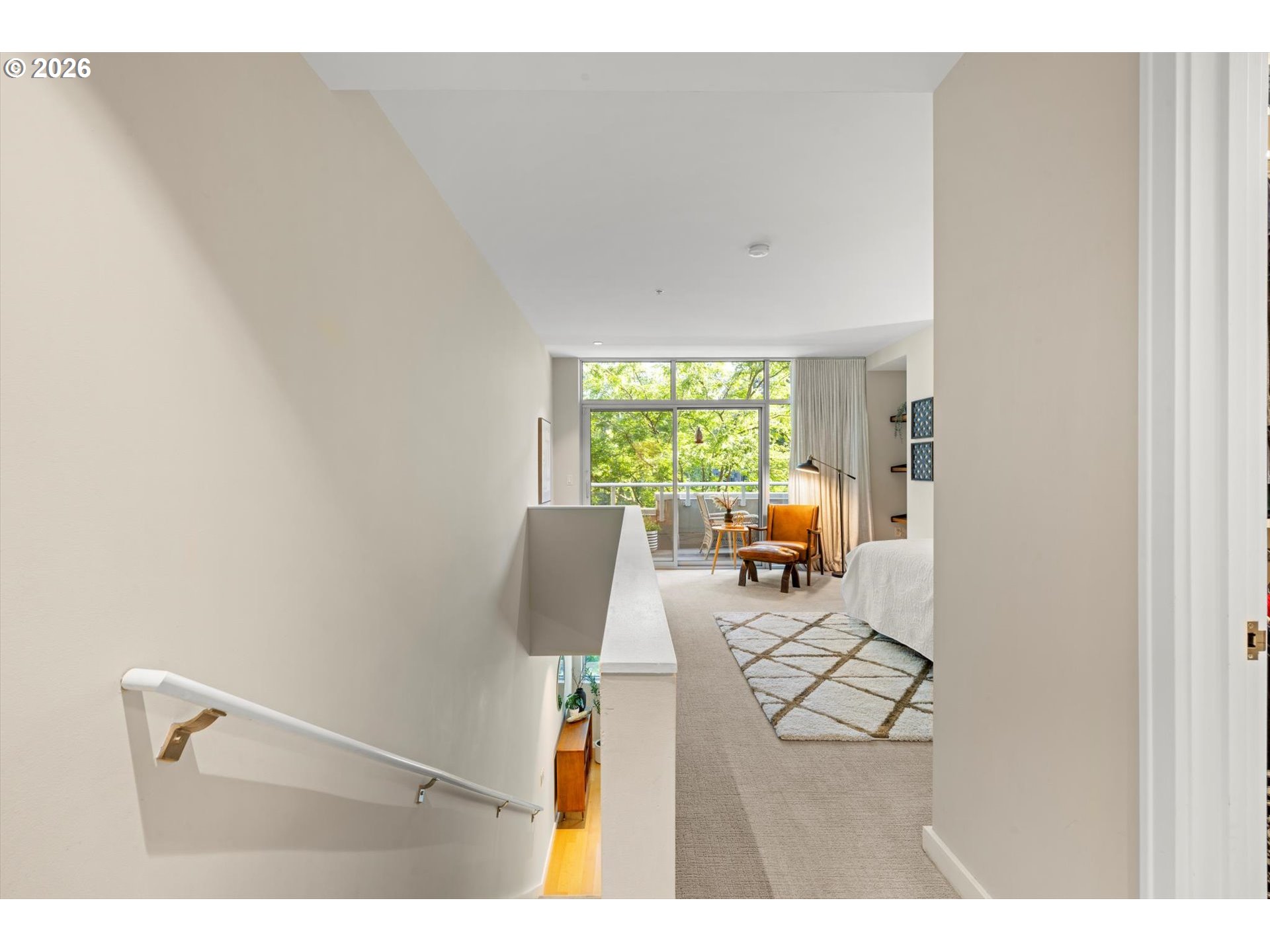 Upstairs landing overlooking a furnished bedroom with a lounge chair and leafy window view.