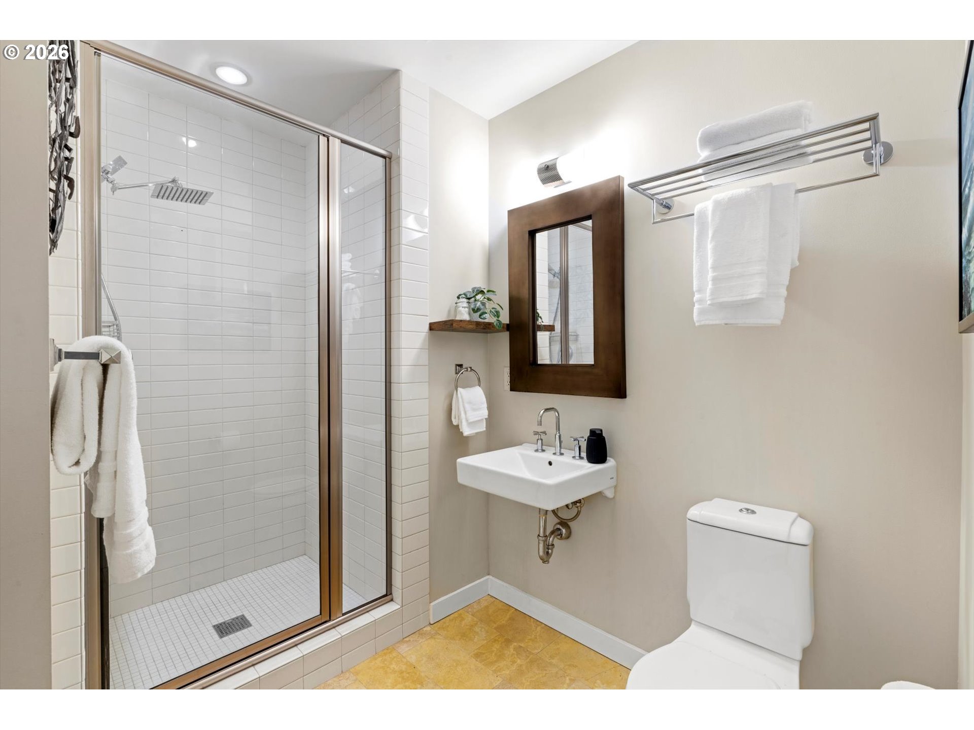 Bathroom with a glass-enclosed tiled shower, wall-mounted sink, dark wood-framed mirror, and golden-toned floor.