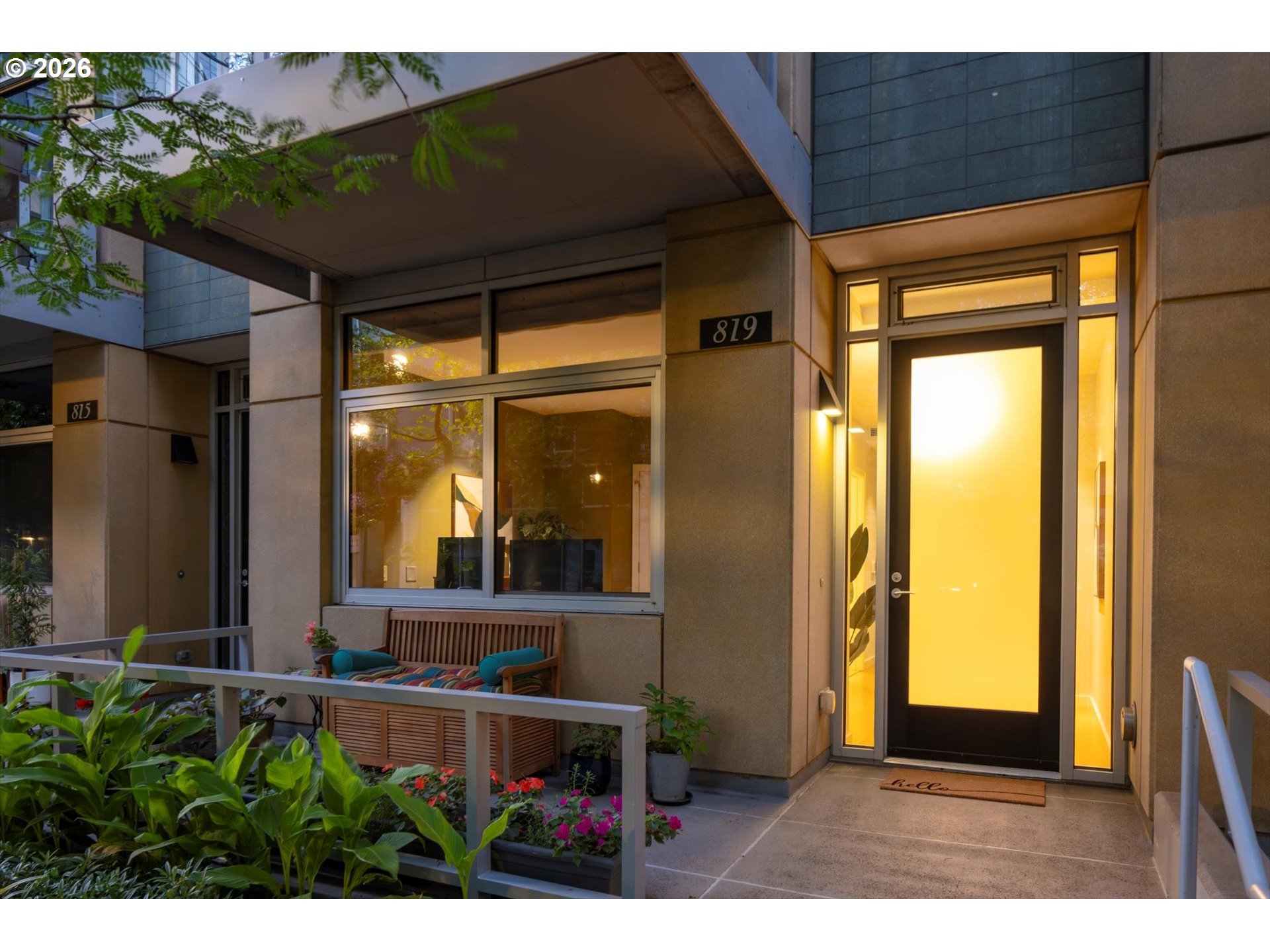 Modern residence entrance with a warmly lit glass door, covered porch, bench, and flower planters.