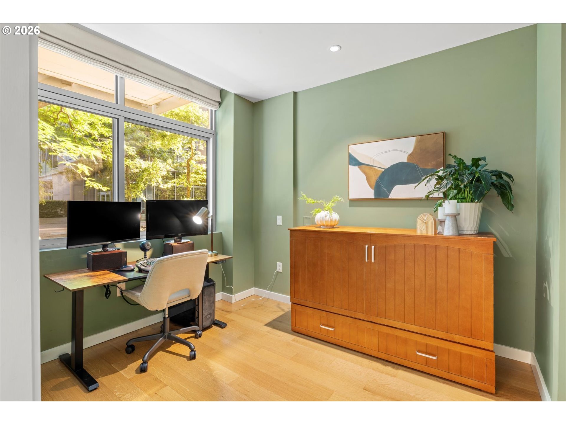 Home office with a dual-monitor desk beside a large window and a wooden storage cabinet.