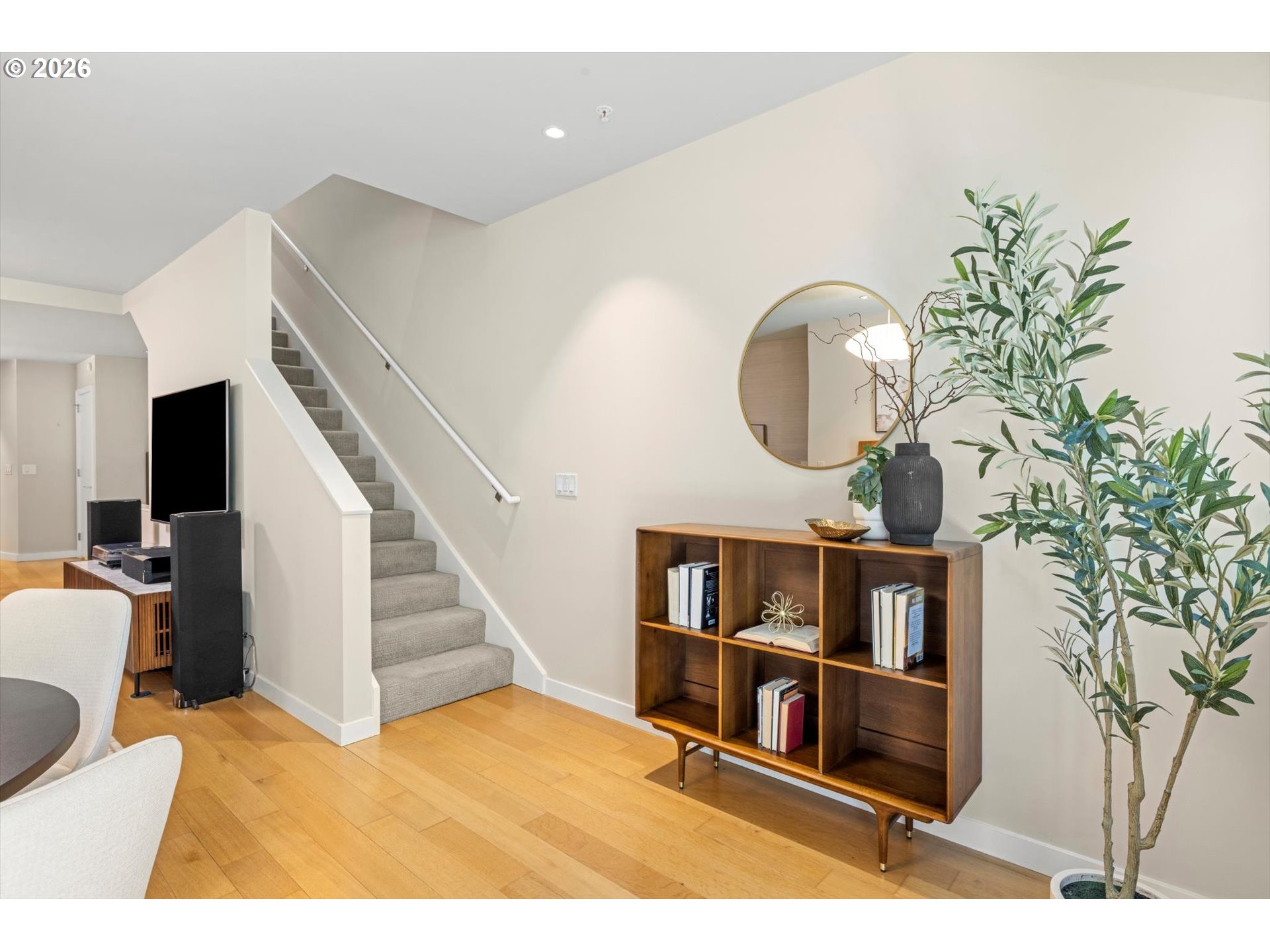 Furnished living area with a carpeted staircase, wooden bookcase, round mirror, and tall potted plant.