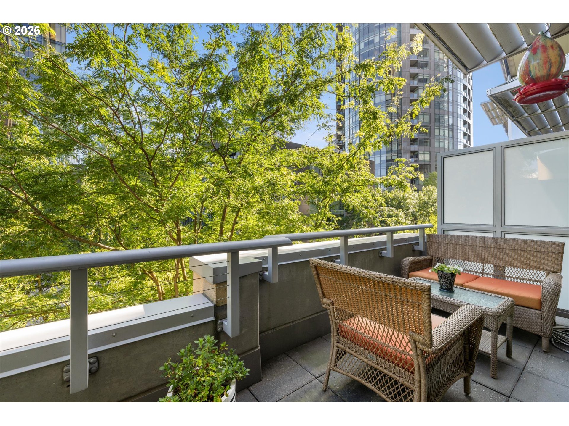 Furnished apartment balcony with wicker seating, leafy trees, and high-rise buildings beyond the railing.