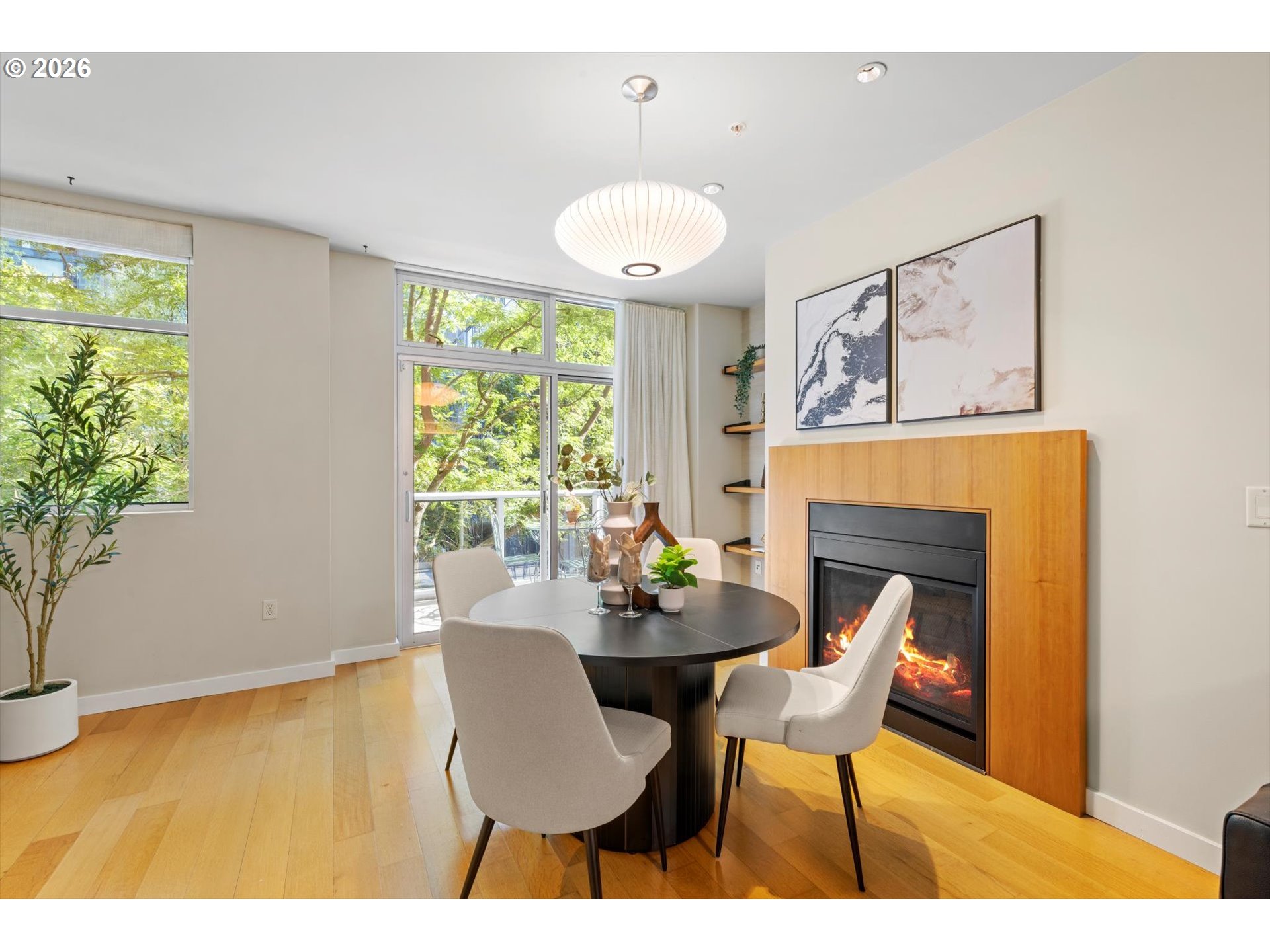 Dining area with a round table beside a lit fireplace and large windows.