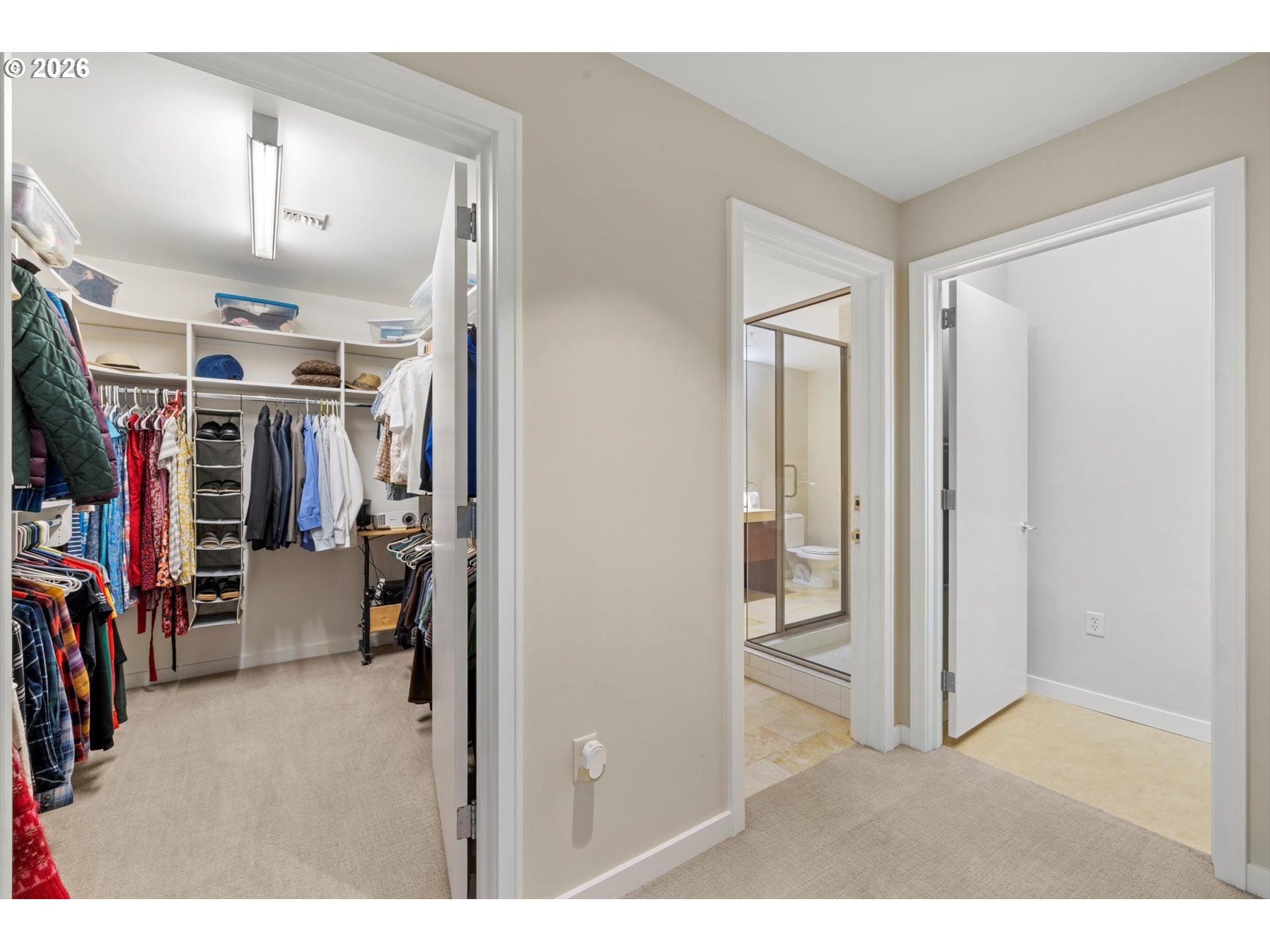 Walk-in closet with built-in shelving and hanging clothes beside a bathroom with a glass shower.