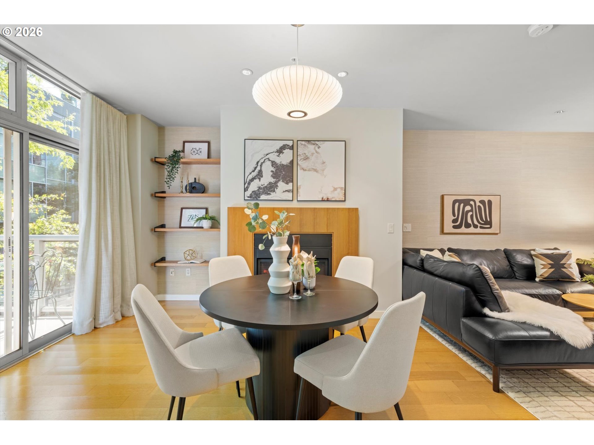 Open-plan living and dining area with a round table, fireplace, black sectional, and large windows.