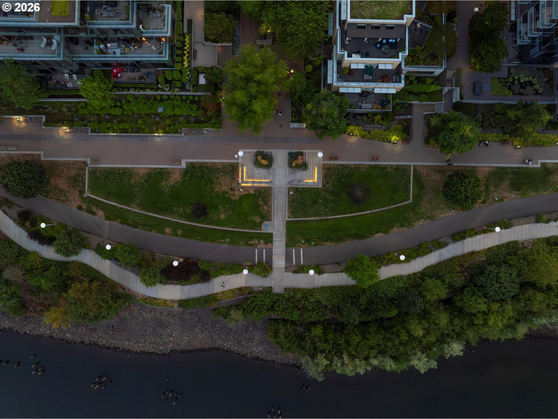 Aerial view of a waterfront promenade with landscaped paths, modern residences, and boats on the river.