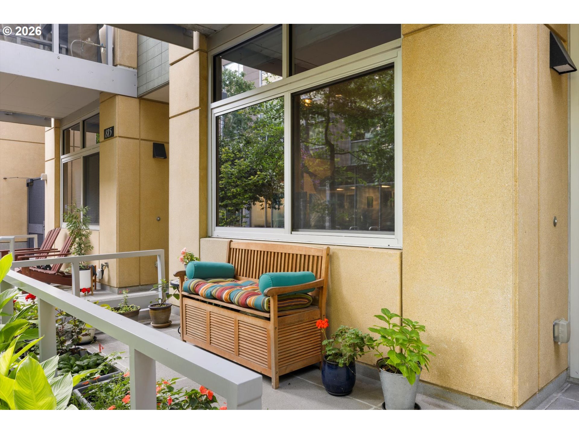 Furnished balcony with a wooden bench, colorful cushions, potted plants, and large windows.