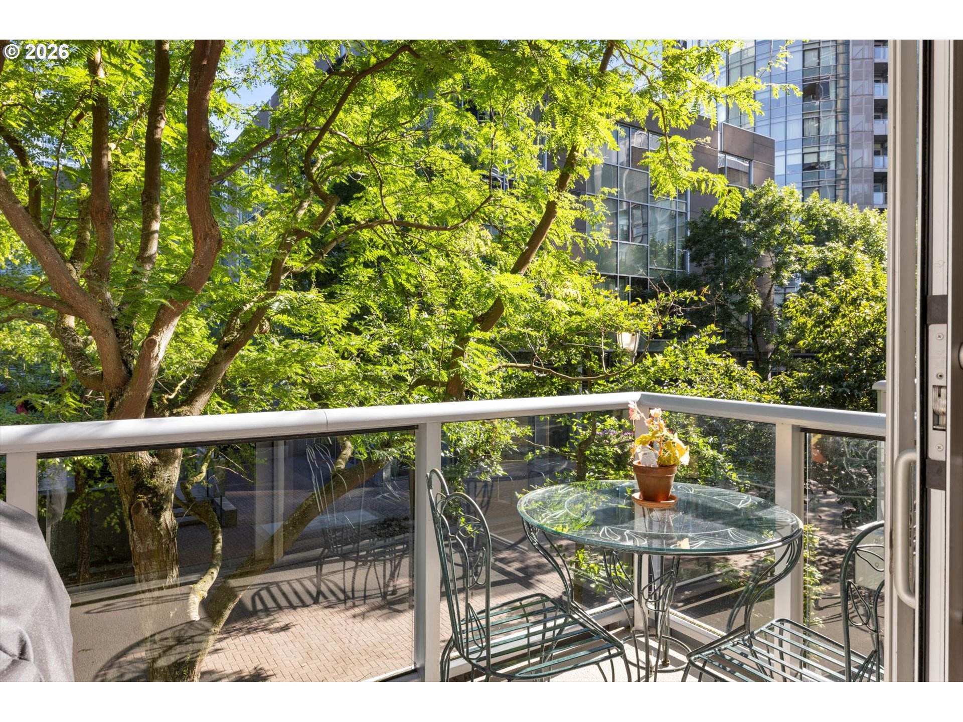 Balcony with a glass table and metal chairs overlooking leafy trees and modern buildings.