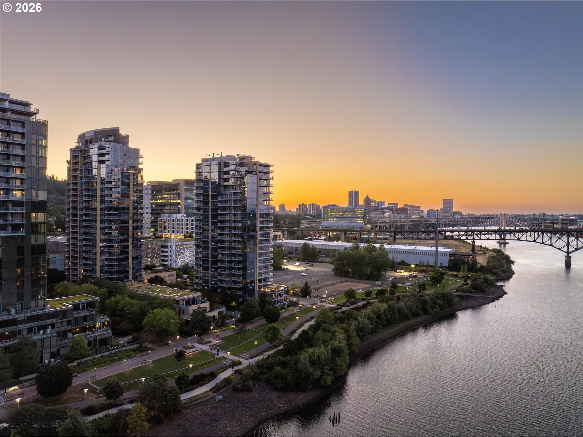 Riverfront city skyline at sunset, with high-rise buildings and a bridge.