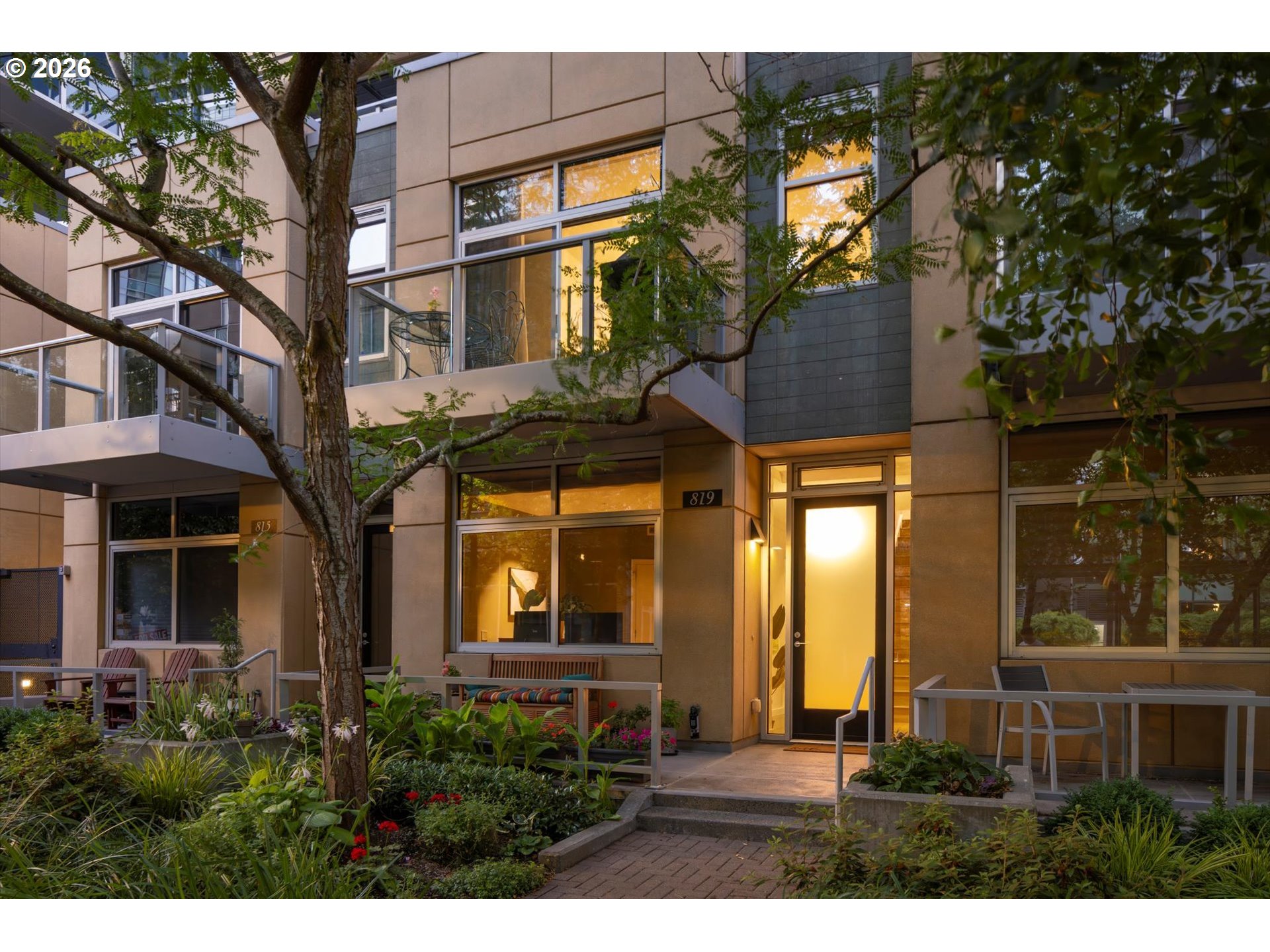 Modern residence entrance with large windows, warm interior lighting, and landscaped gardens.