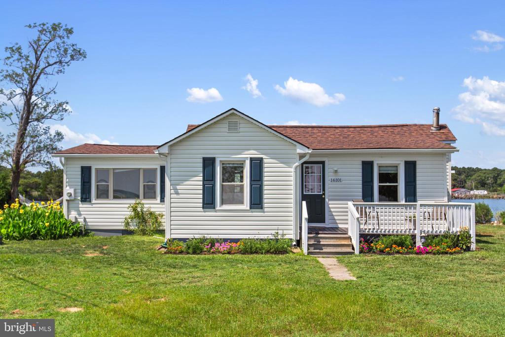 Single-story light-sided home with dark shutters, a white-railed porch, flower beds, and water nearby.