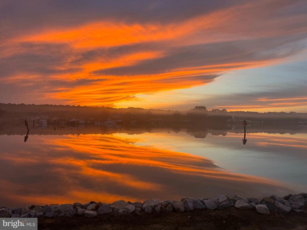 Calm waterfront reflects a vivid orange sunset, with boats across the water and rocks along the shore.