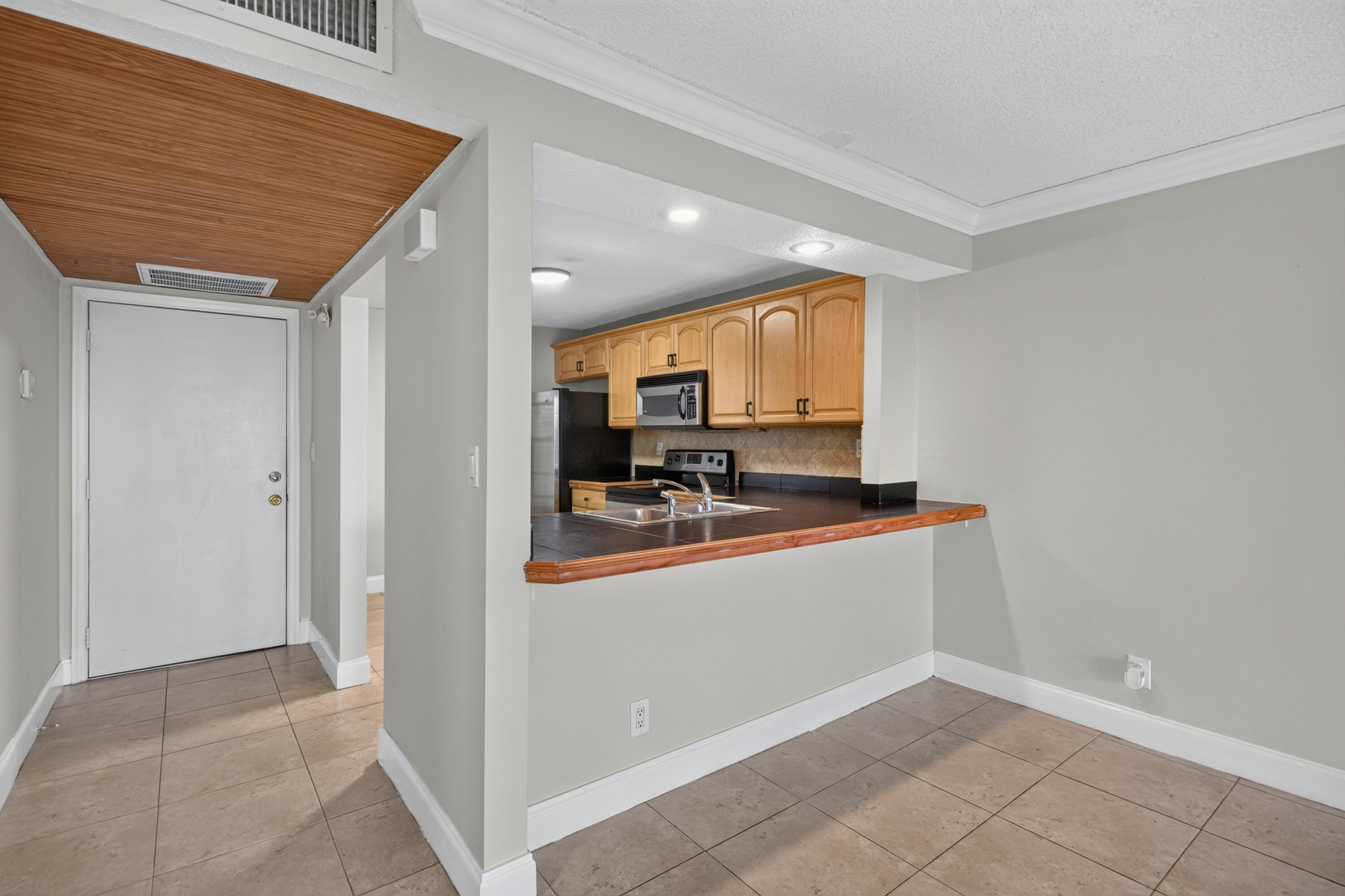 Condo kitchen and entry with honey-toned cabinets, dark breakfast bar, and tan tile floors.