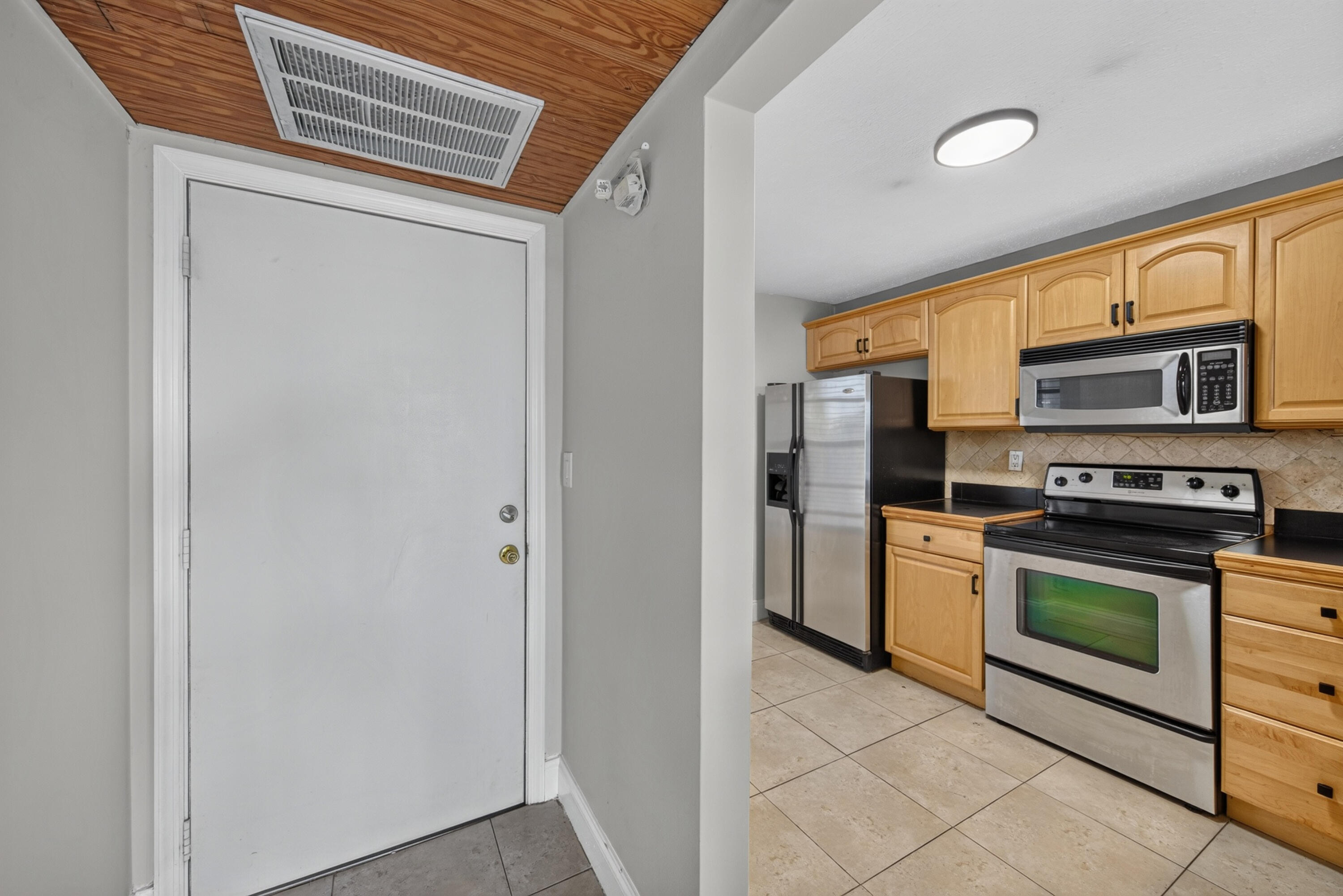 Entryway beside a kitchen with light wood cabinets, stainless steel appliances, and a wood-paneled ceiling.