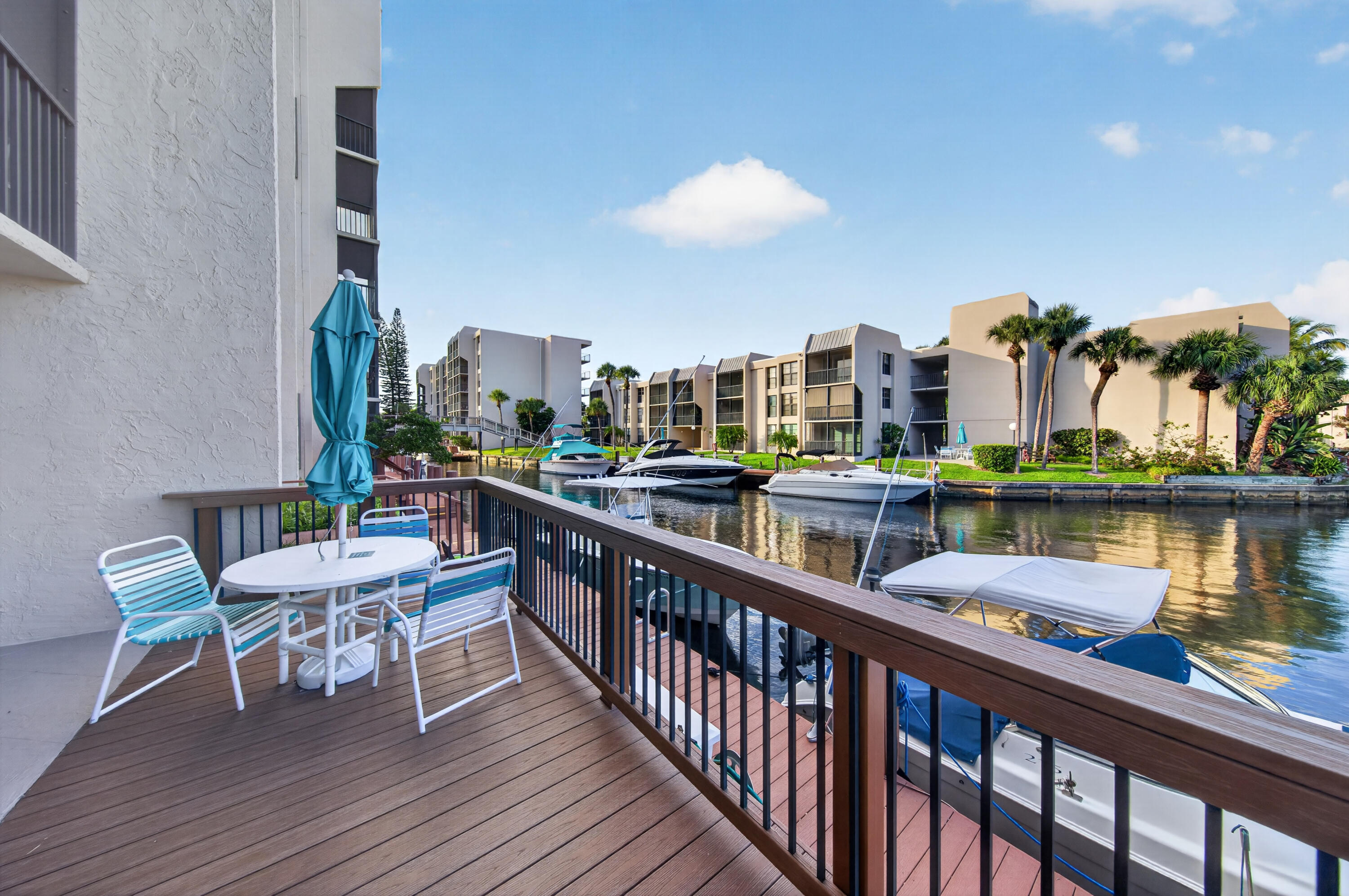 Condo balcony overlooking a canal, with striped chairs, a turquoise umbrella, and boats moored nearby.