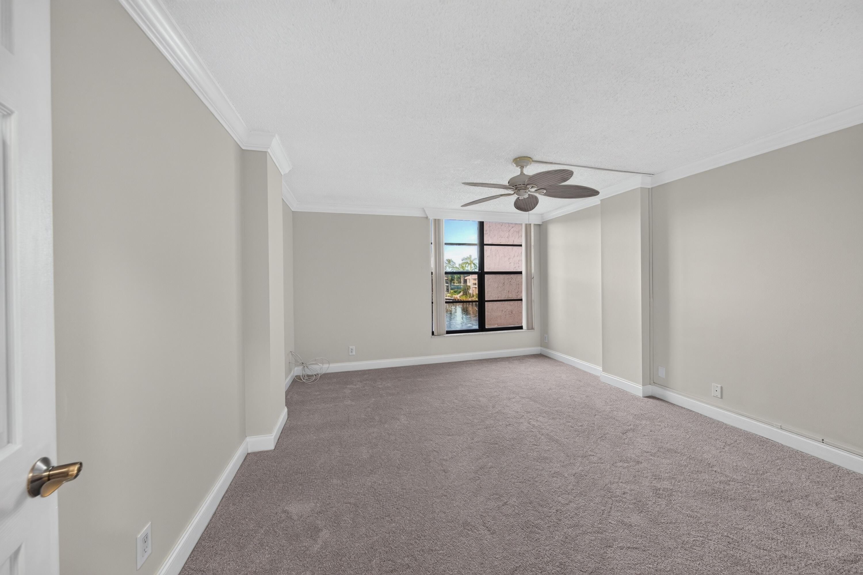 Empty carpeted room with a ceiling fan and a window overlooking water and palm trees.