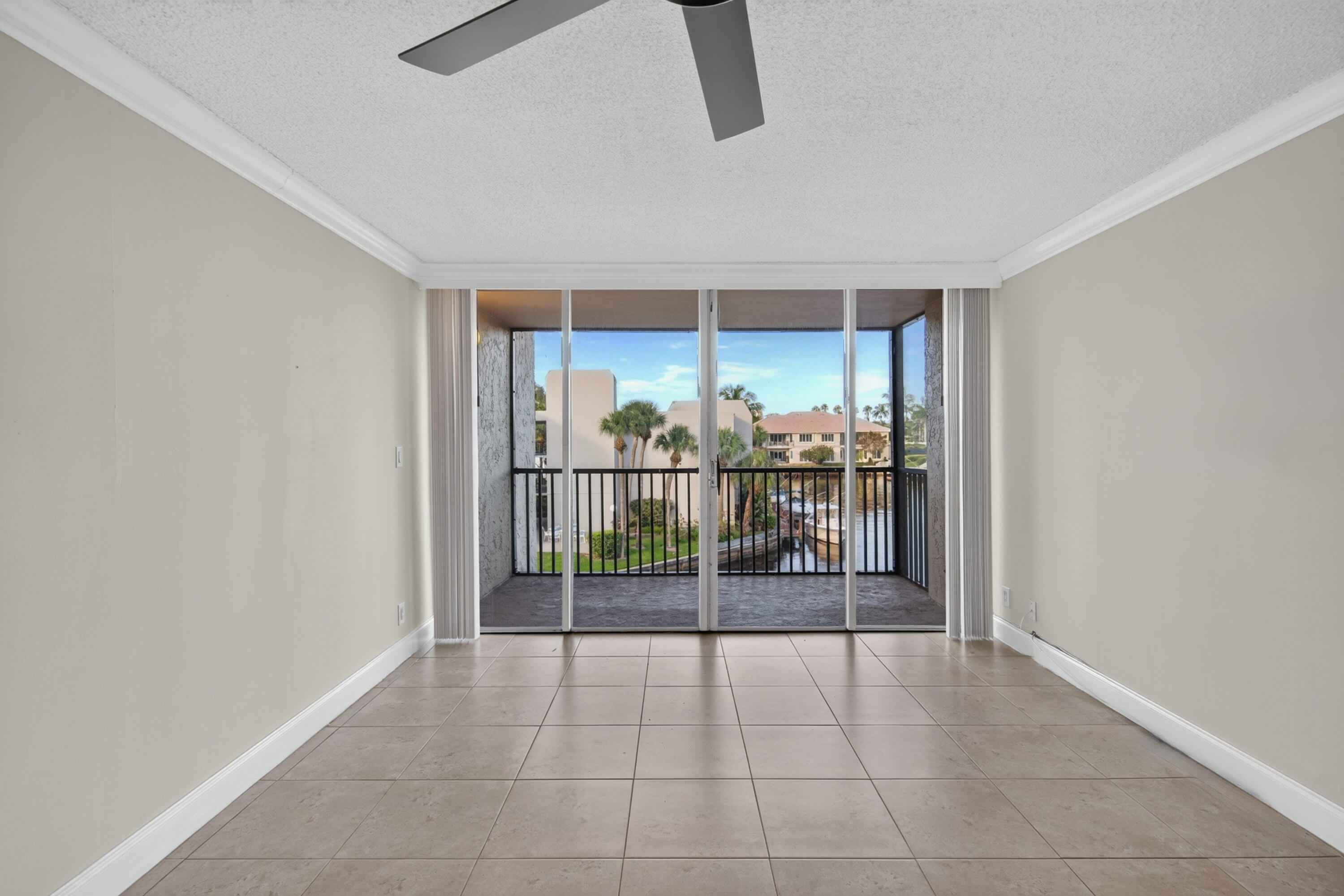 Empty tiled room with sliding doors to a balcony overlooking palms, neighboring homes, and a canal.