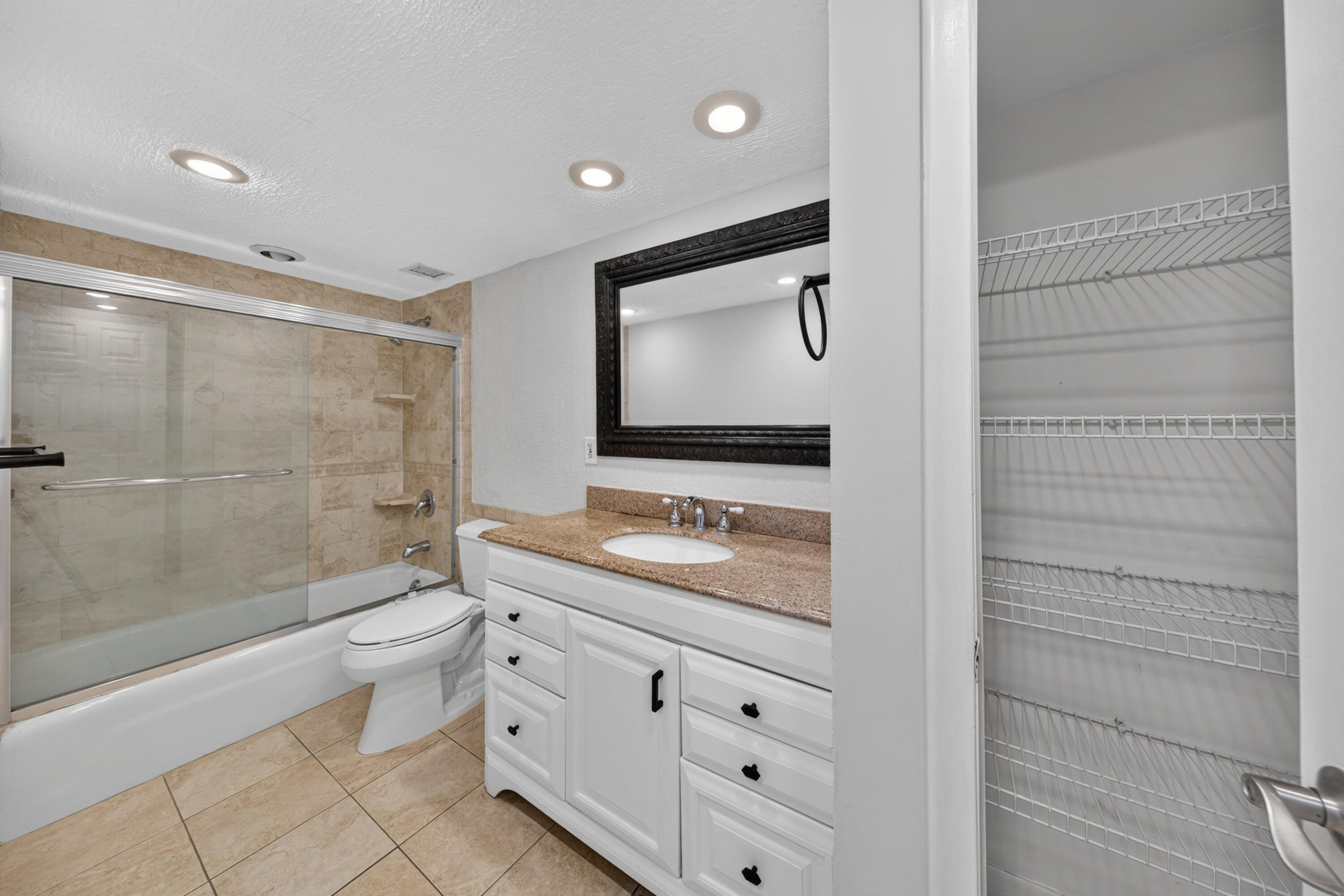 Bathroom with a glass-enclosed tub, granite-topped vanity, and open wire-shelf closet.