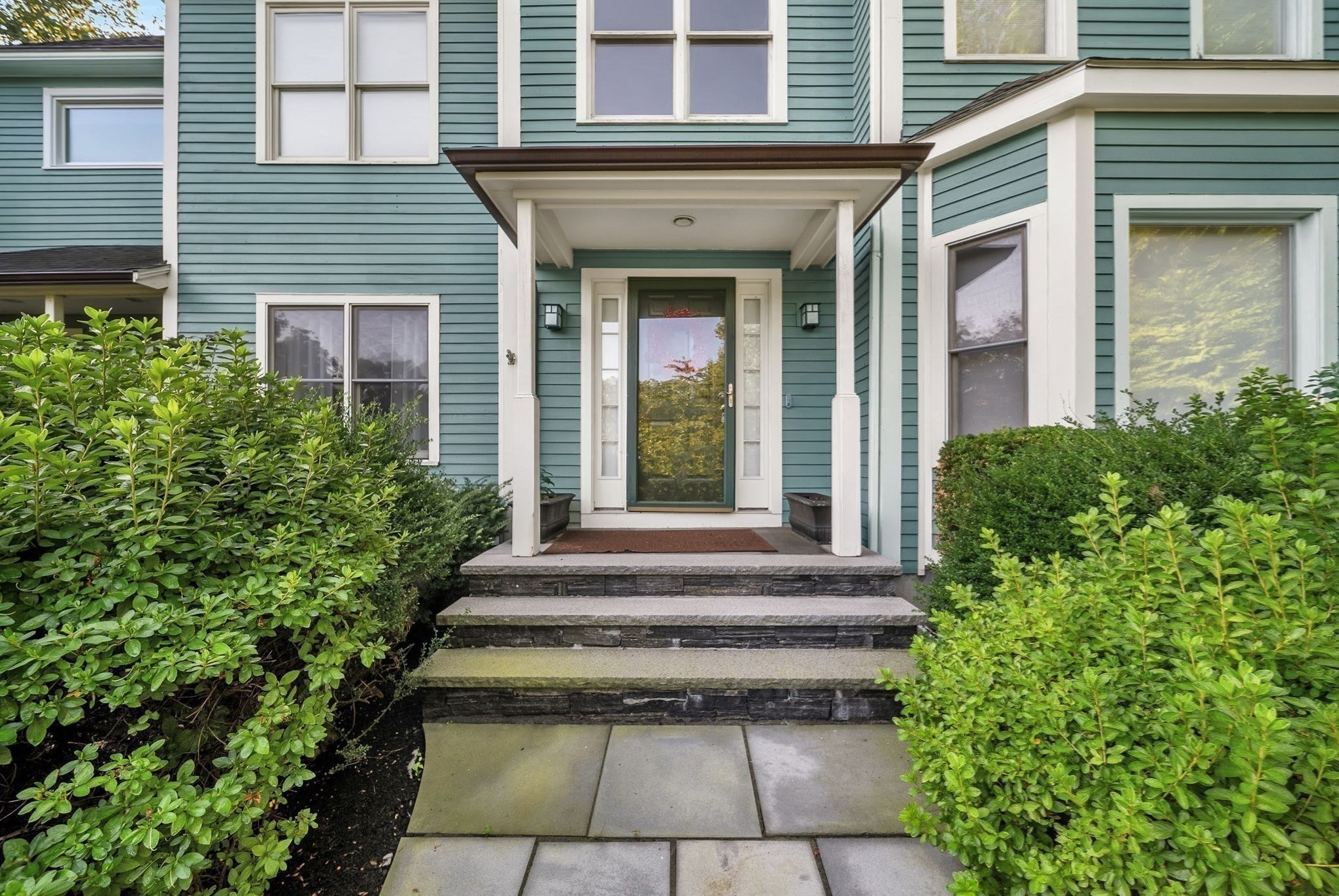 Blue-green house entrance with a covered porch, stone steps, and dense shrubs.