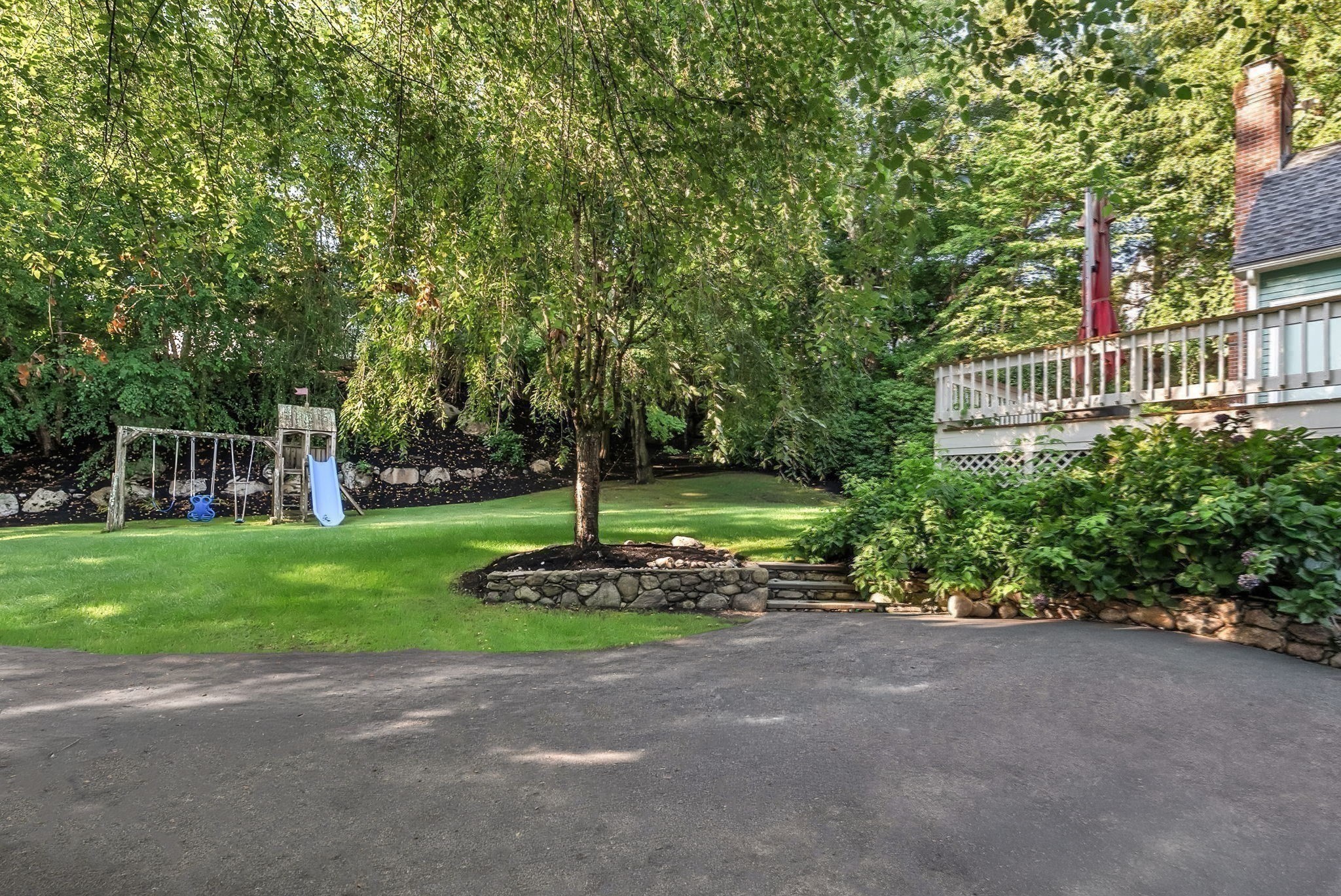 Tree-shaded backyard with a lawn, children's play set, stone retaining wall, and raised deck.