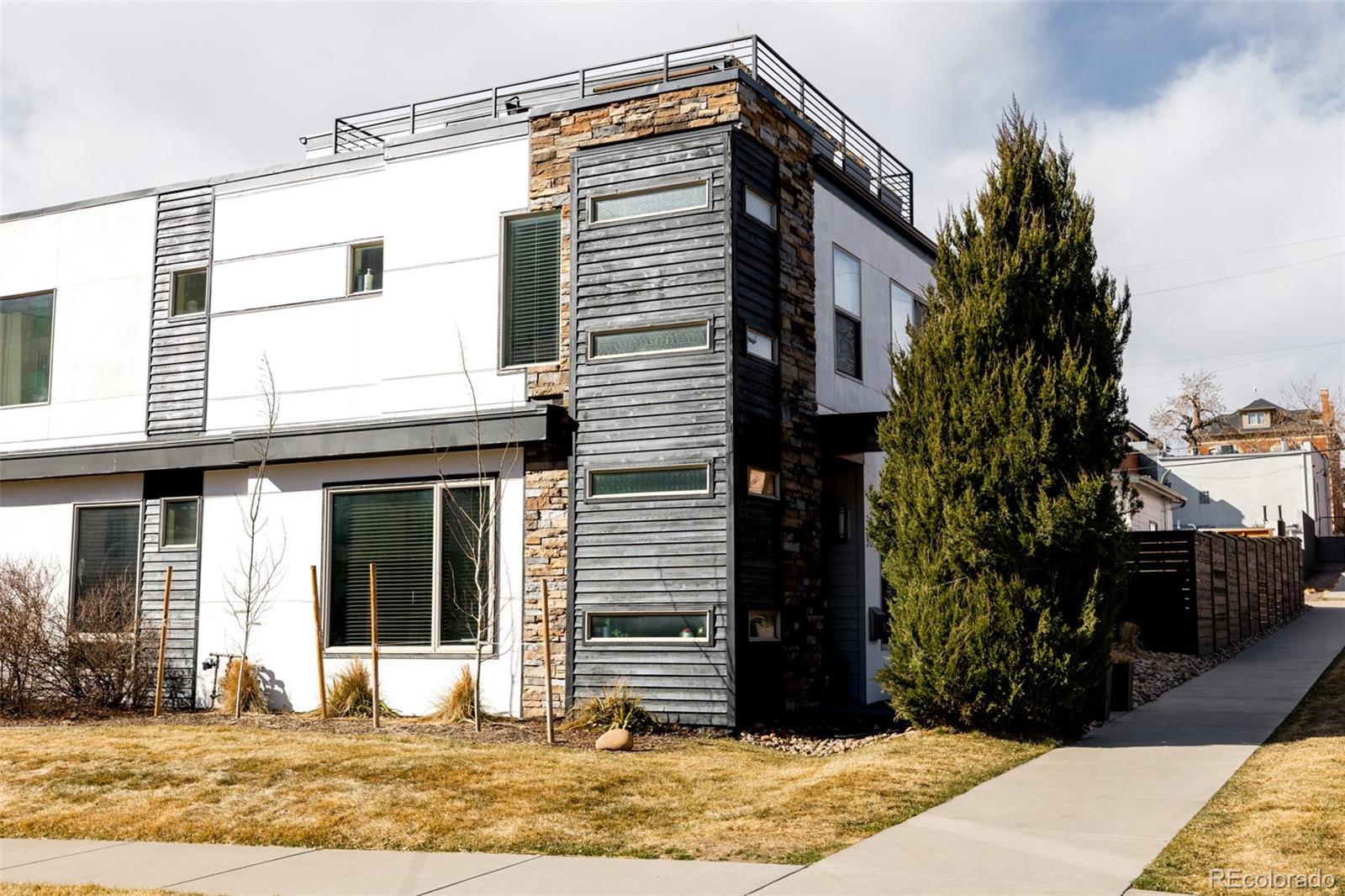 Modern white and gray home with stone accents, rooftop terrace railing, and a tall evergreen by the sidewalk.