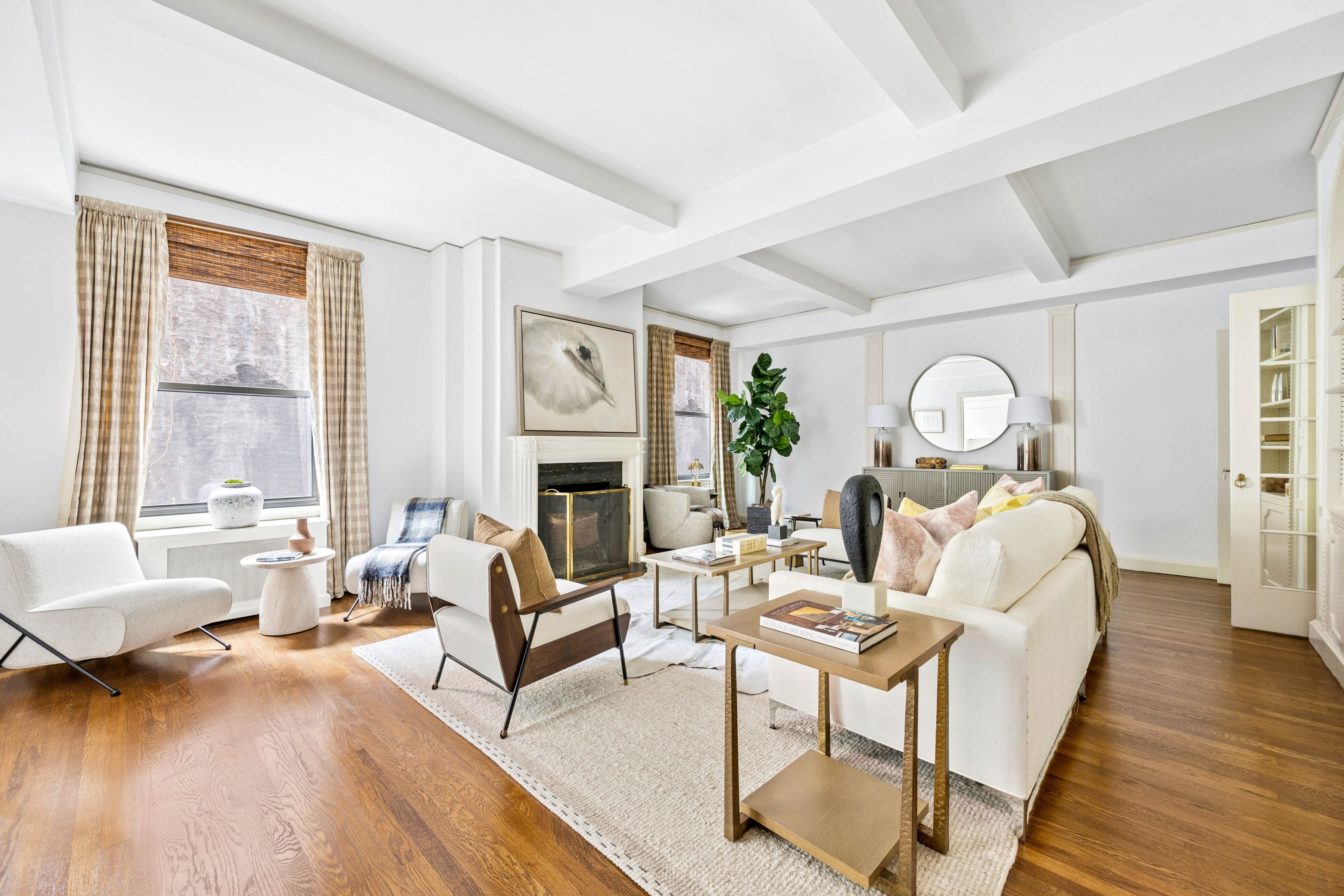 Bright living room with a fireplace, beamed ceiling, hardwood floors, and white seating.